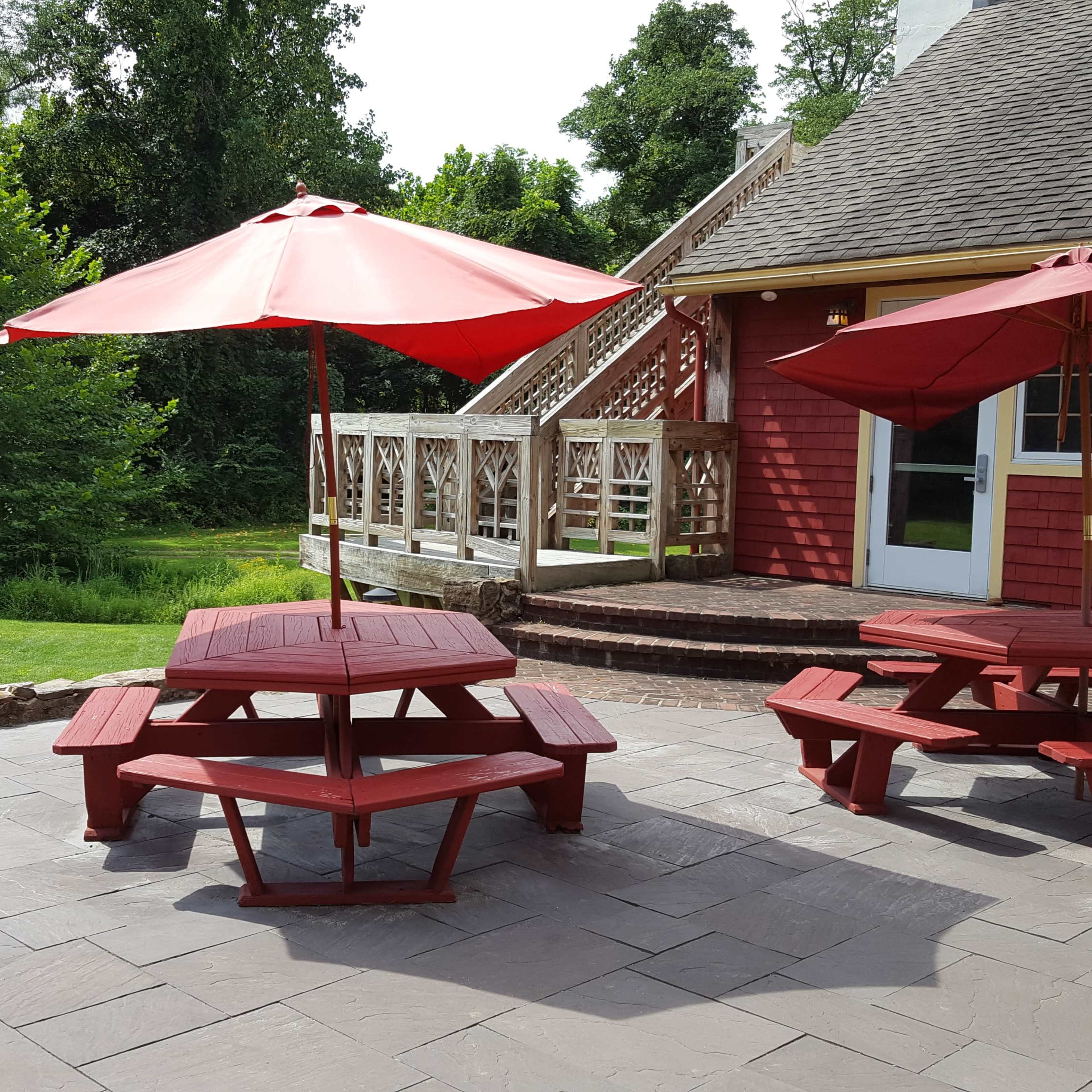 A patio area with two red picnic tables under matching umbrellas, surrounded by a green lawn and a staircase leading to the upper level of a building.