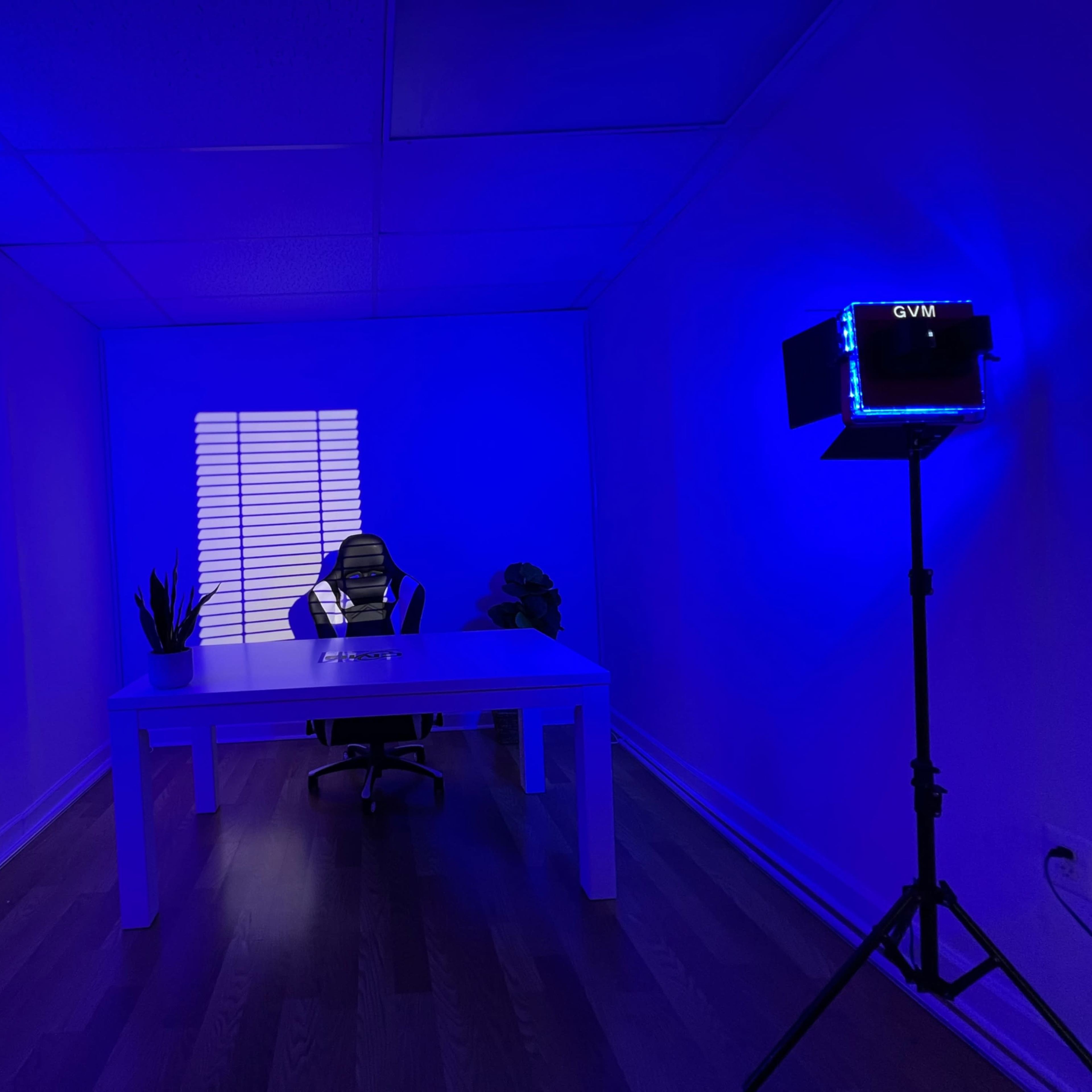 An empty room with a white desk and chair, illuminated by blue lighting from two standing lamps, with a window casting shadows on the wall.