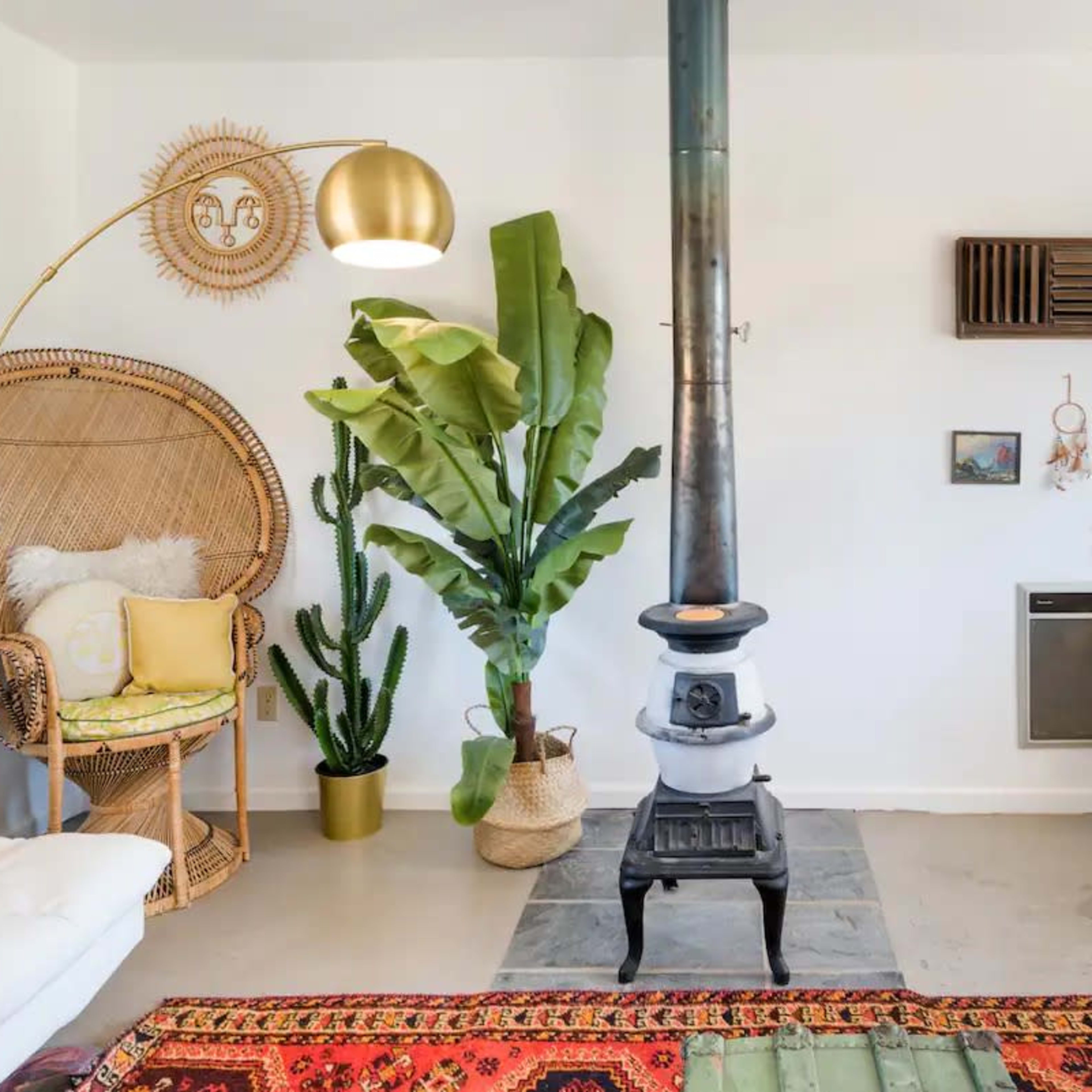 A cozy living room features a wood-burning stove, an ornate floor lamp, various plants, and decorative wall hangings.