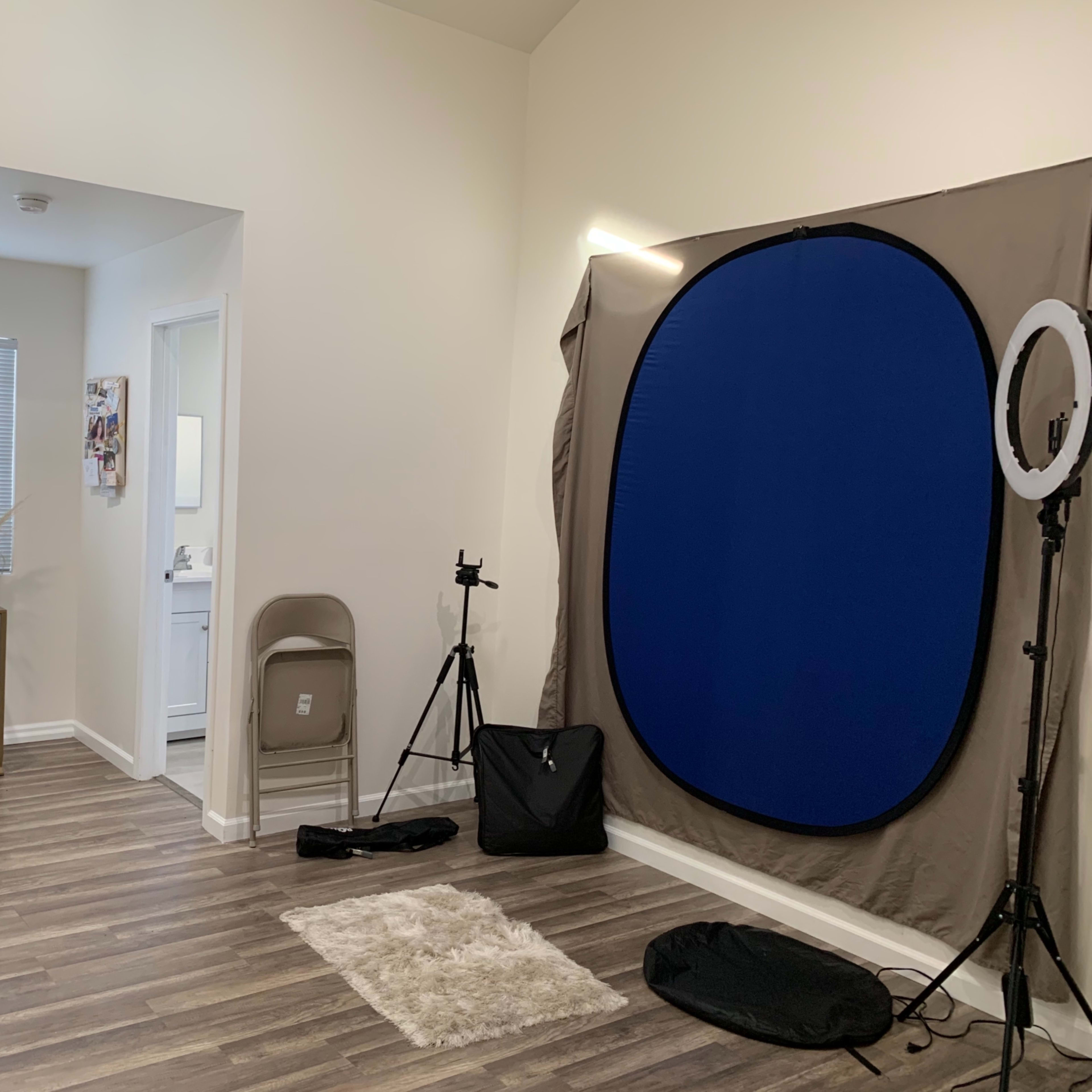 A well-lit room featuring a small table and chair, a photography backdrop, a ring light, and a decorative vase in the corner.