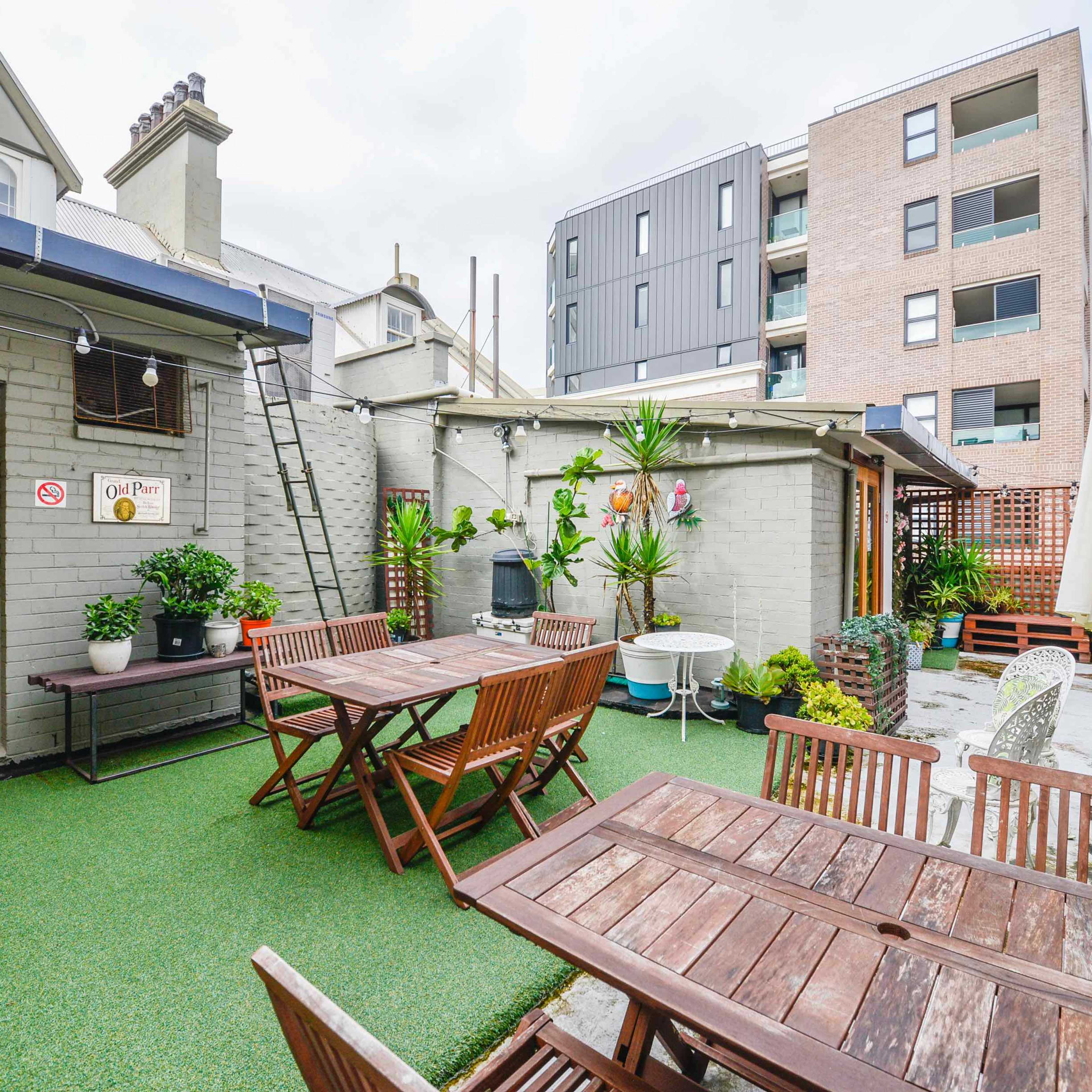 An outdoor patio area with wooden tables and chairs, surrounded by plants and modern buildings in the background.