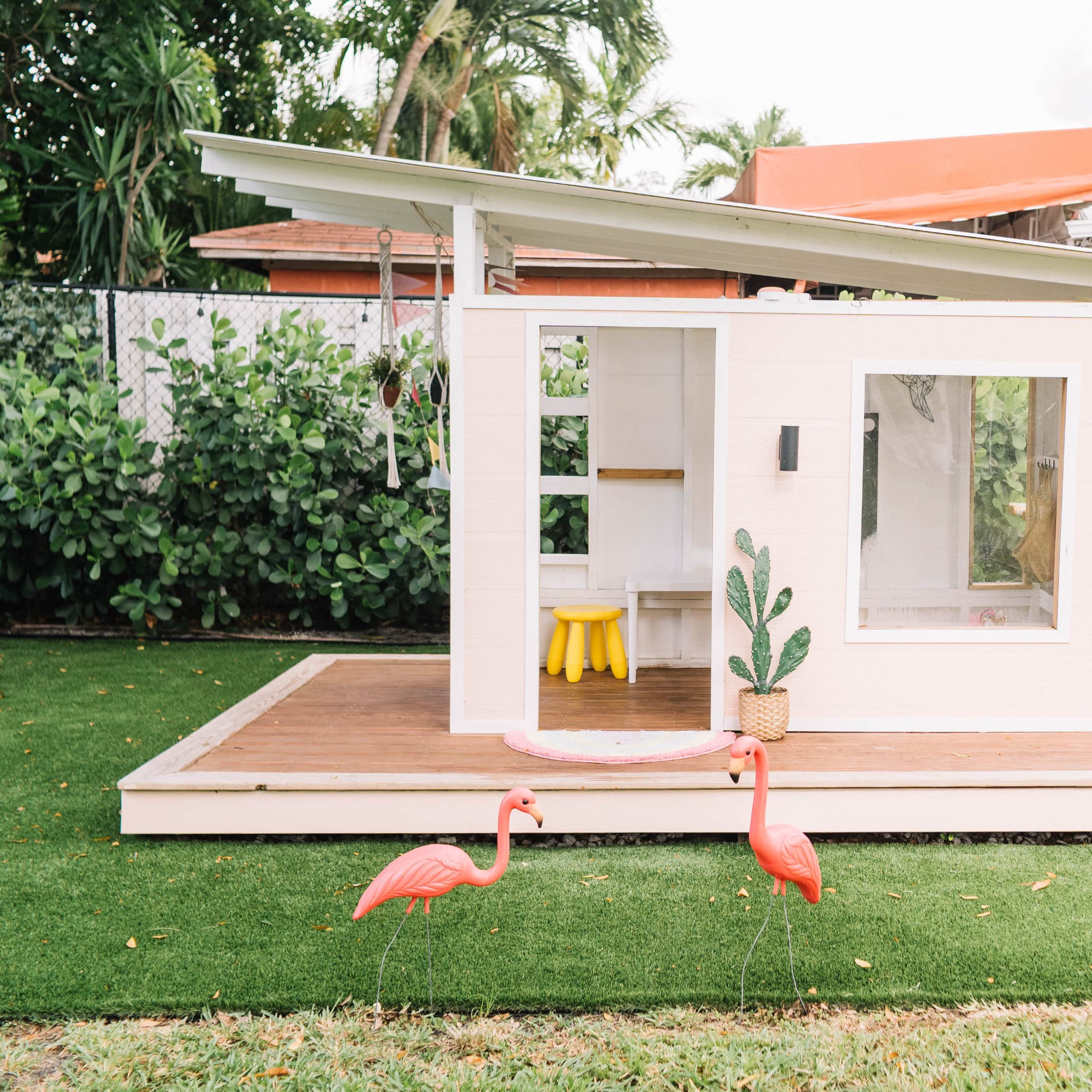 A small, modern playhouse with a slanted roof is situated on a grassy area, accompanied by two pink flamingo lawn decorations.