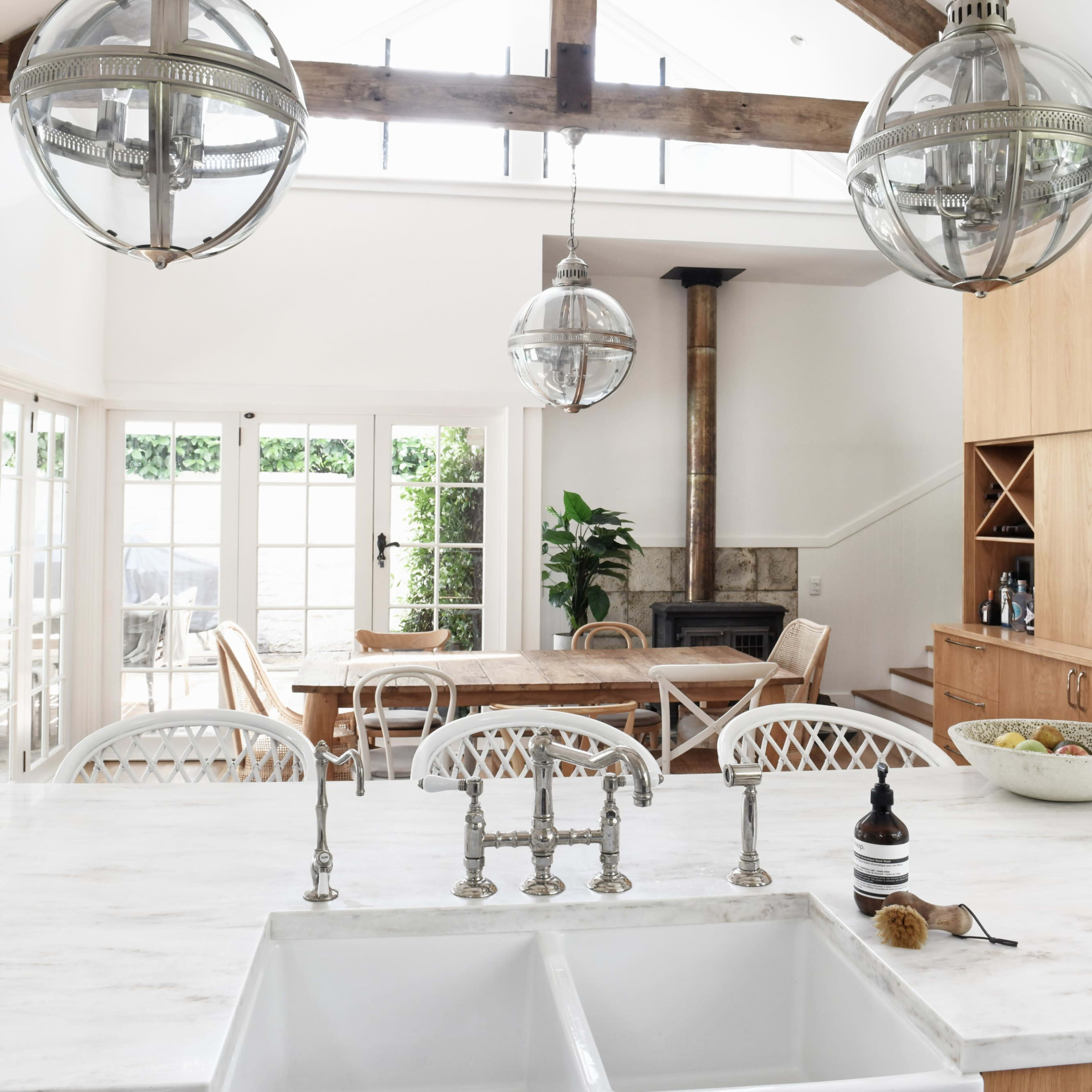 A bright kitchen with a large farmhouse sink, a wooden dining table in the background, and three pendant lights hanging from a vaulted ceiling.