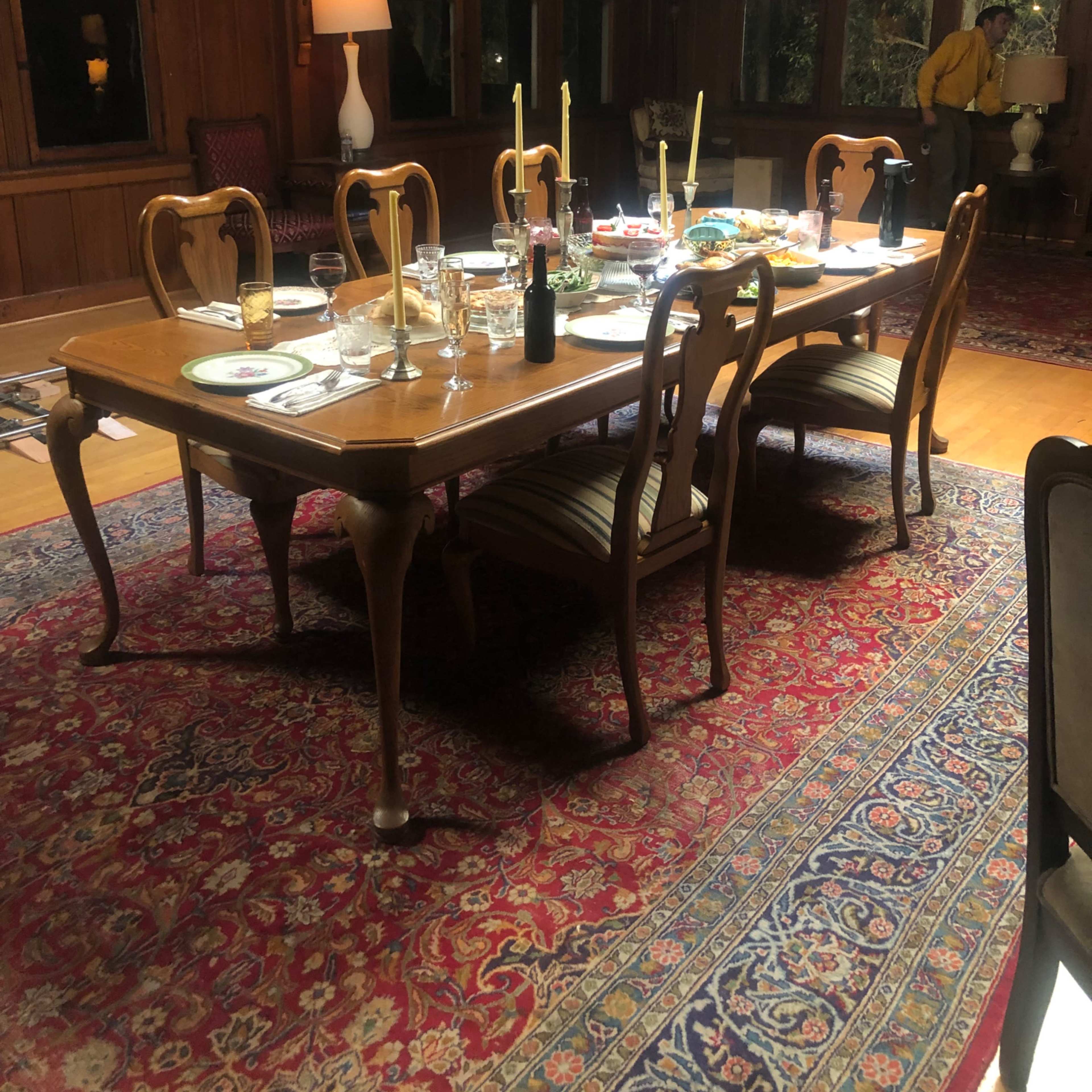 A wooden dining table is set with dishes and candles in a warmly lit room featuring large windows and wooden paneling.