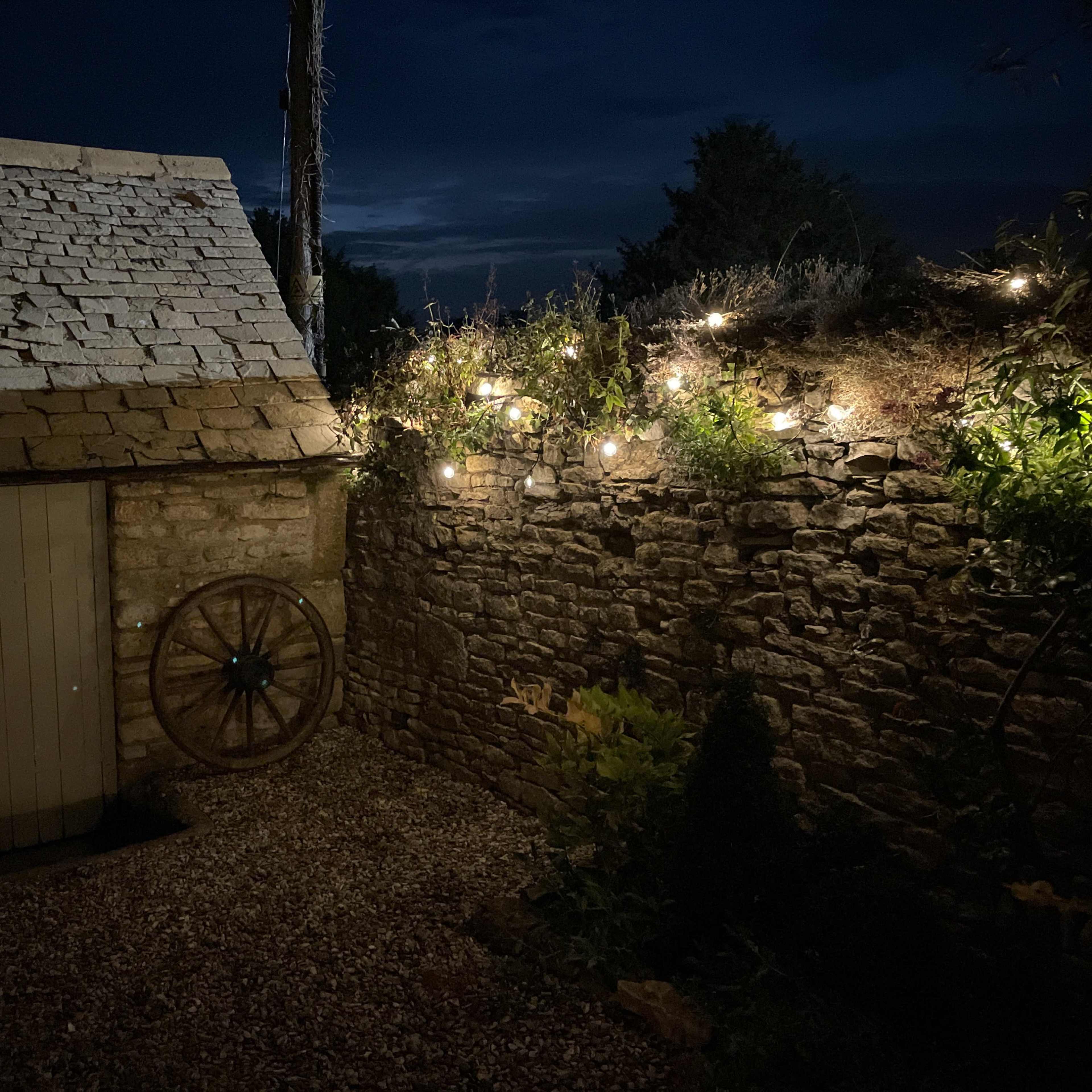 A small stone building with a slanted roof sits beside a lit stone wall adorned with string lights at dusk.