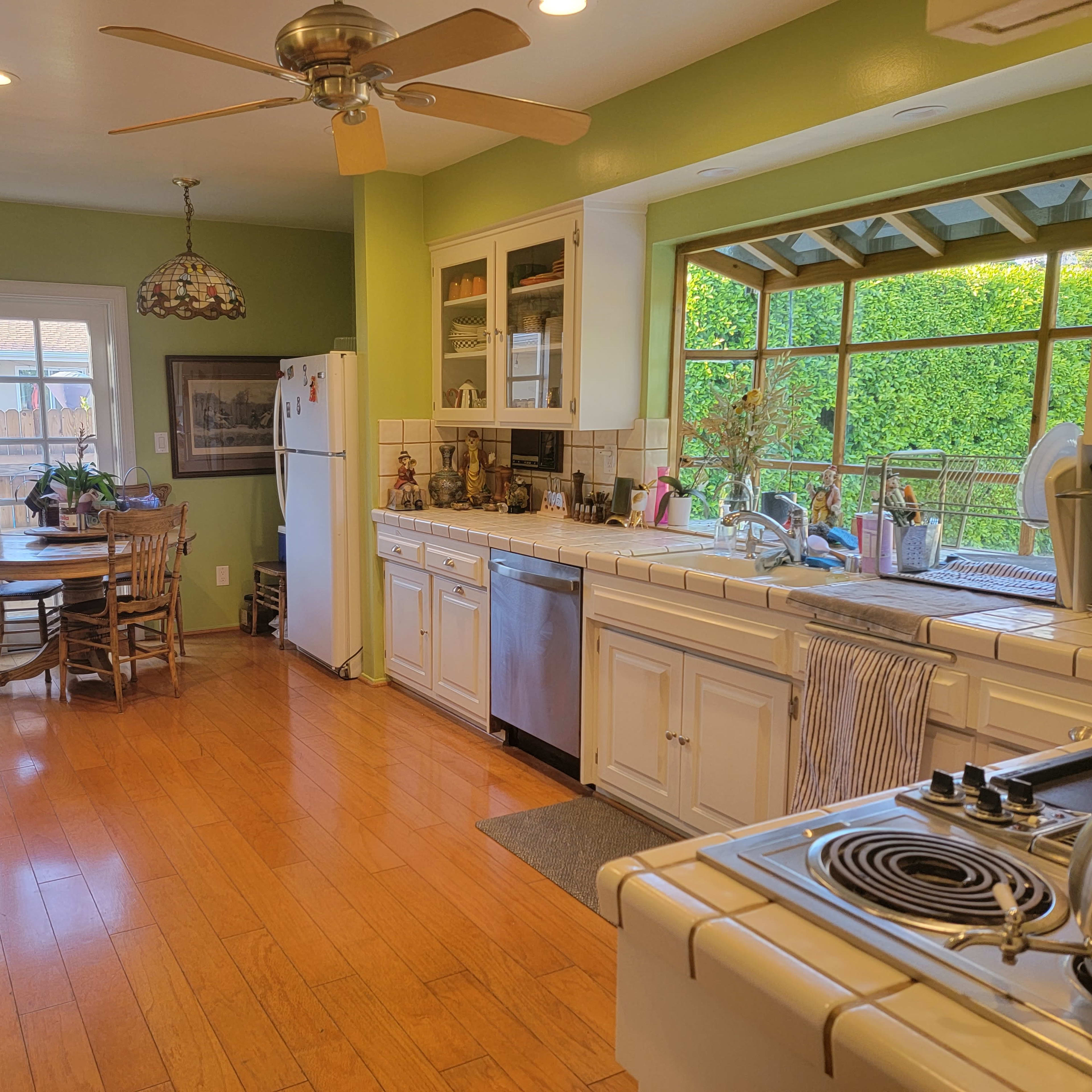 A bright kitchen features white cabinetry, a central island with a stove, and an eating area with a round table and chairs, all illuminated by natural light from a large window.