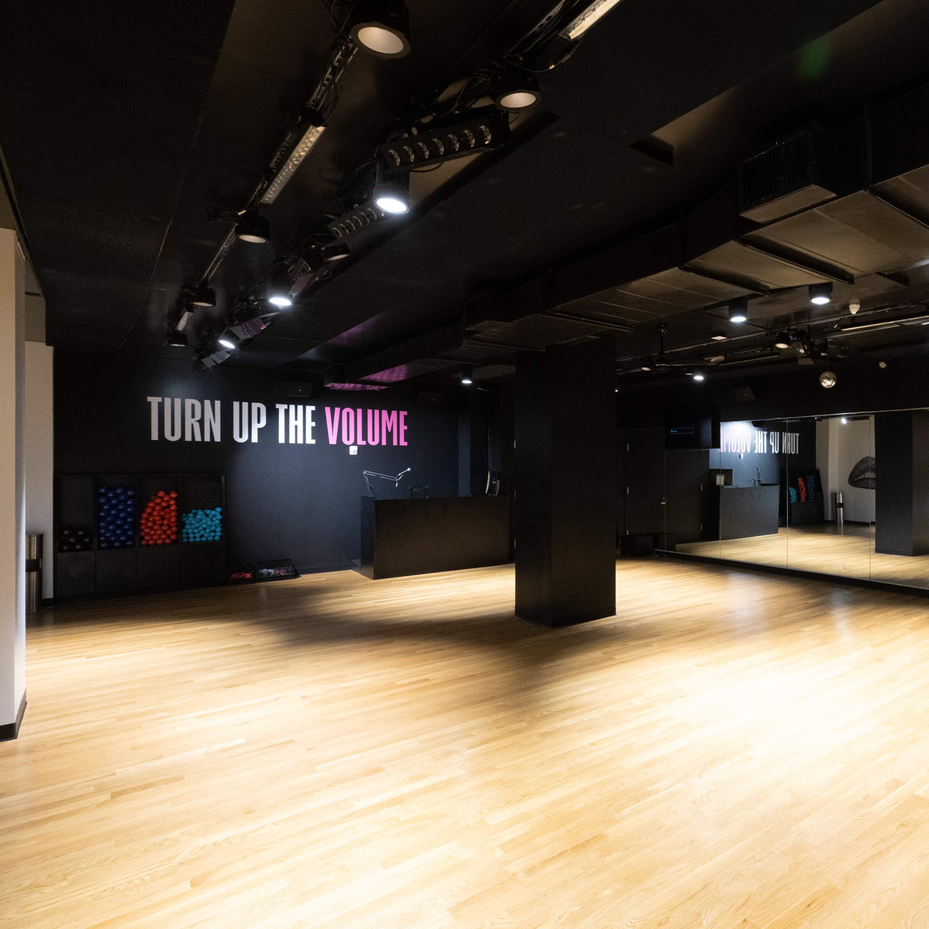 A spacious, modern studio with wooden flooring, black walls, and large mirrors, featuring a "TURN UP THE VOLUME" sign.