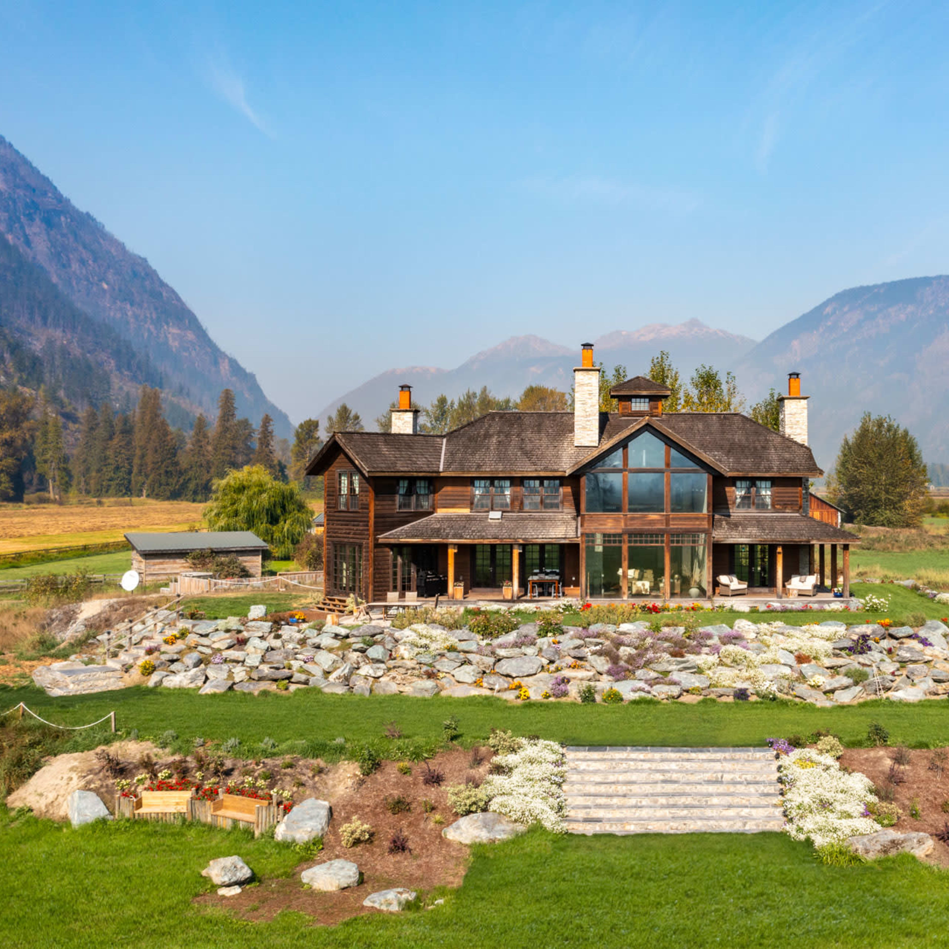 A large wooden house with a stone foundation is situated on a grassy lawn surrounded by mountains, with a landscaped stone pathway leading to the entrance.