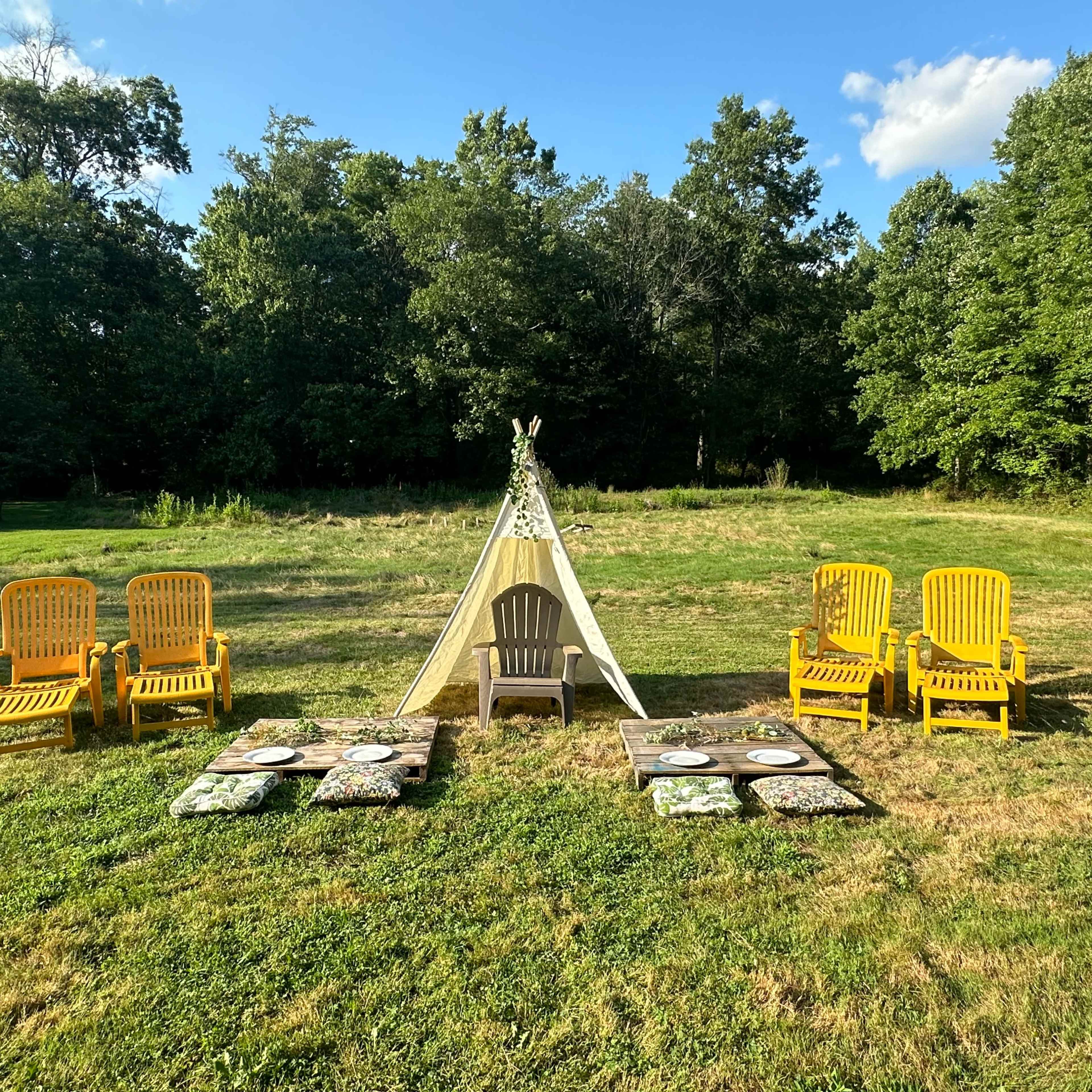 A teepee stands in a grassy area surrounded by yellow chairs and decorative plates arranged on the ground.
