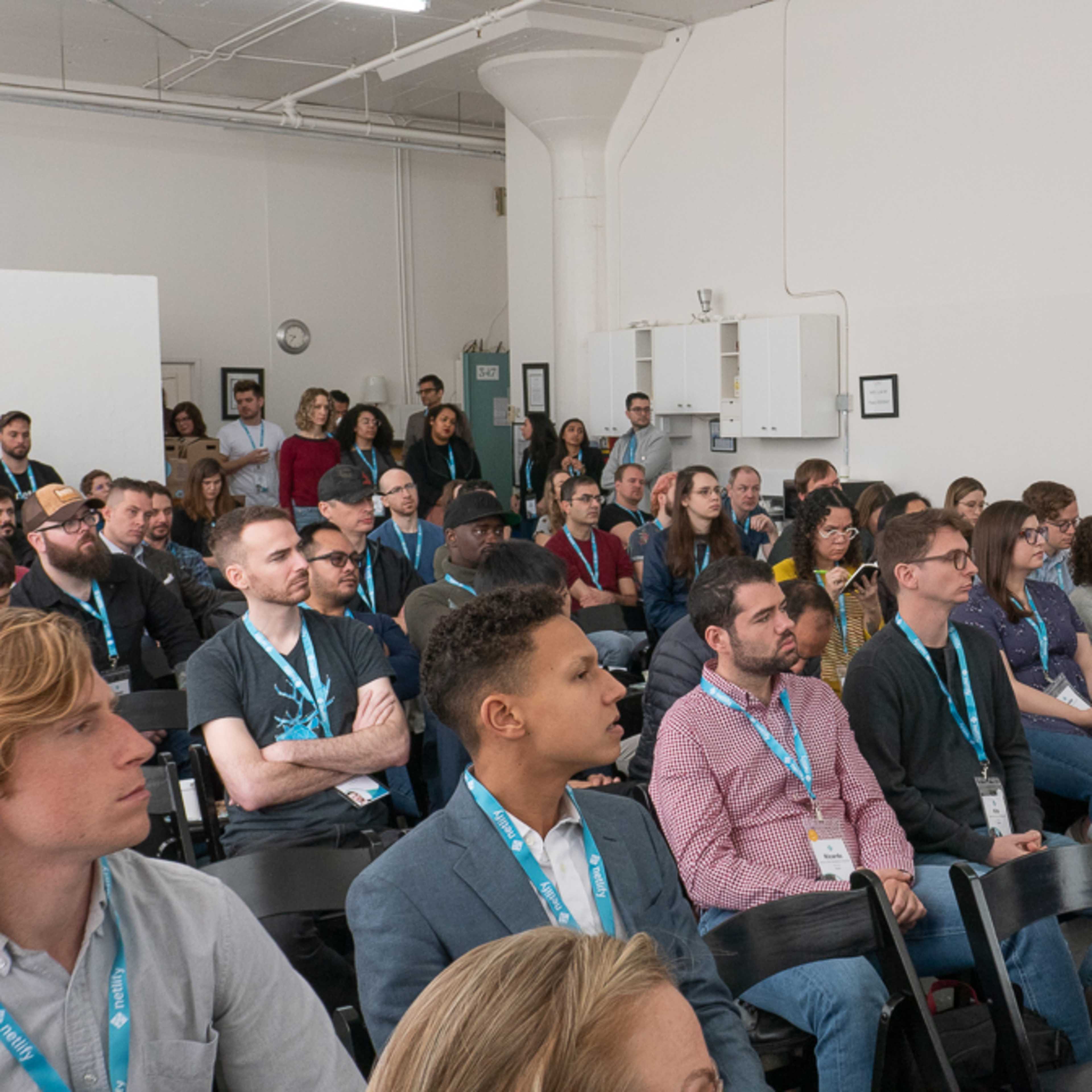 A diverse audience sits in rows of chairs, attentively facing a speaker in a brightly lit conference room.