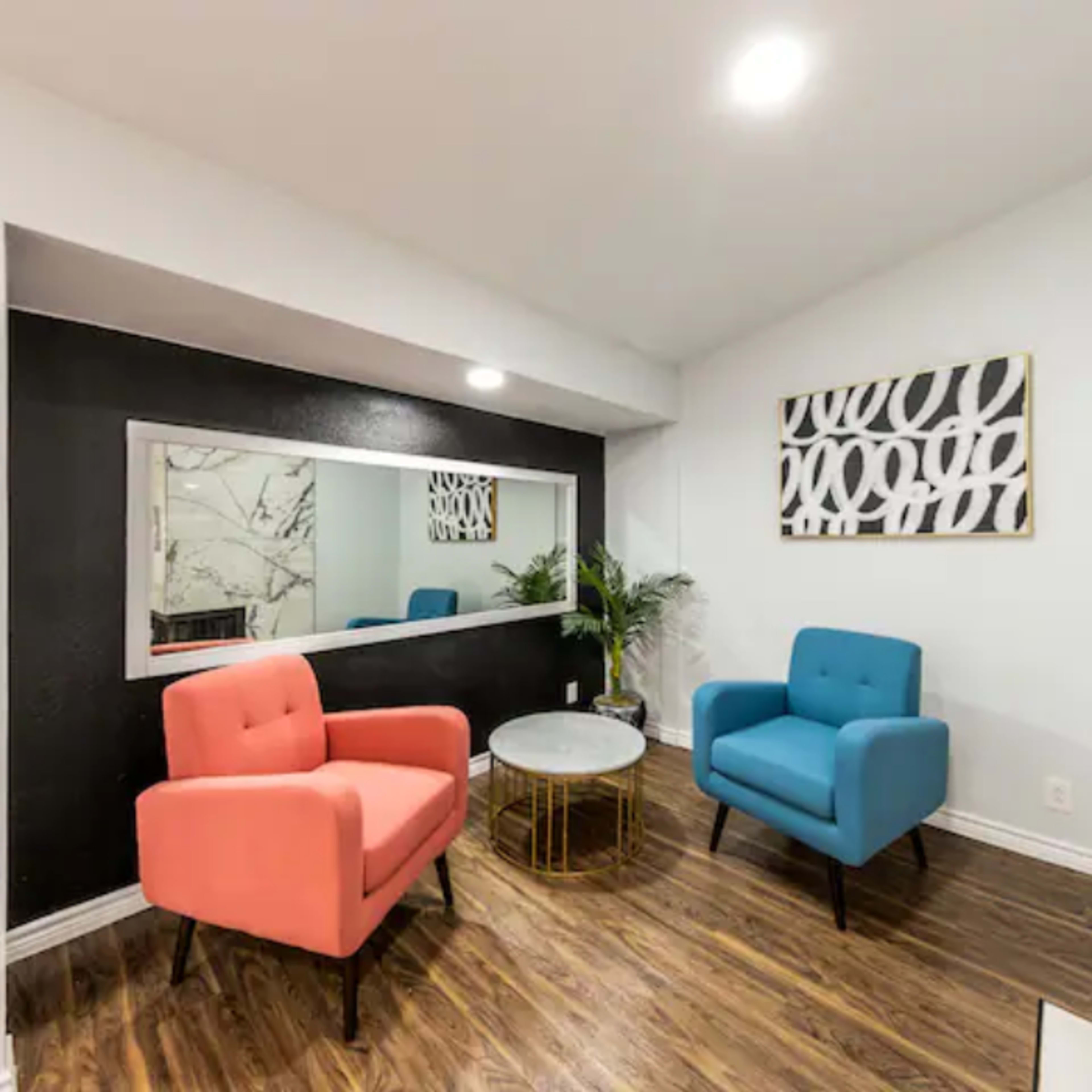 A small room featuring two colored chairs, a round table, a large mirror, and a piece of wall art.