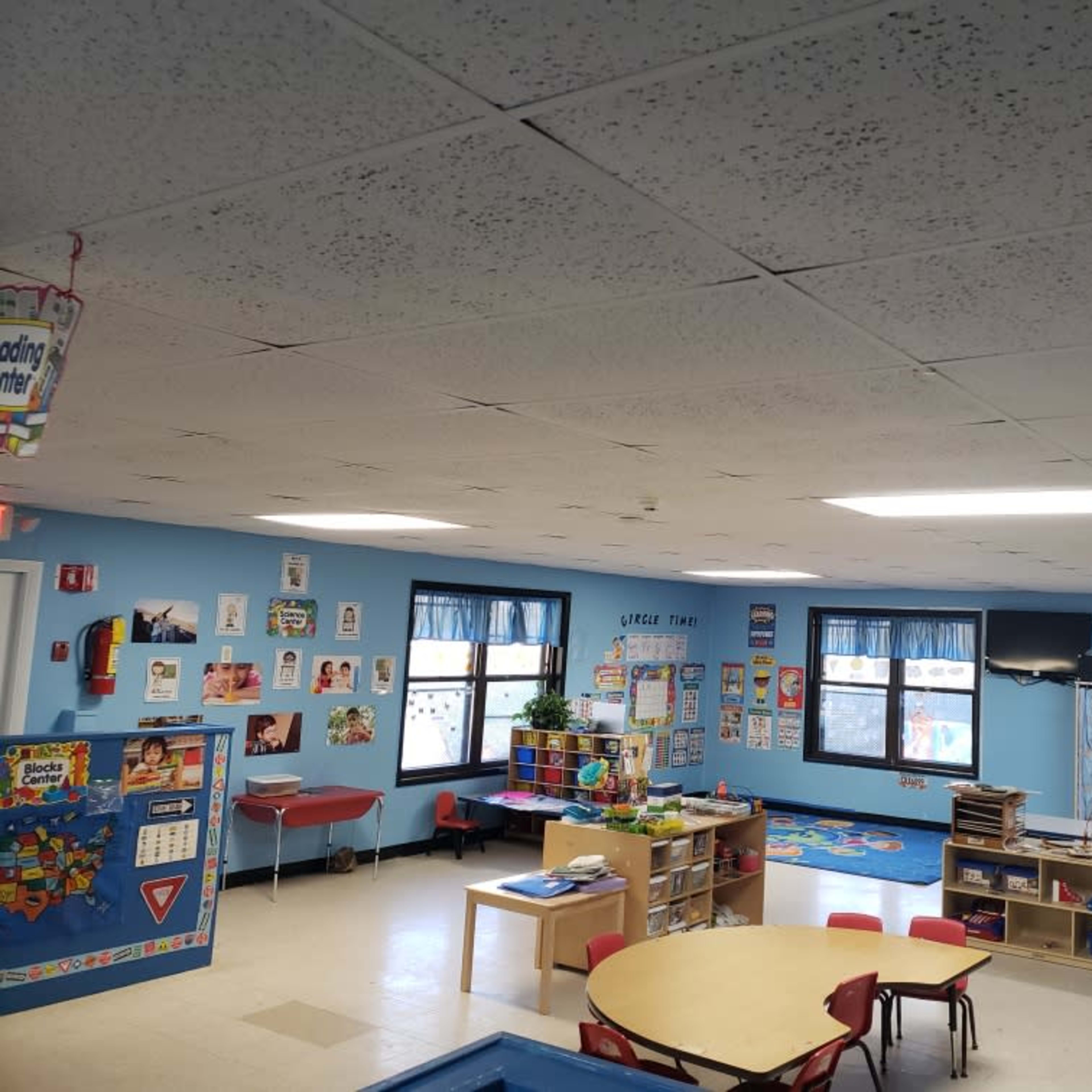 A brightly lit classroom features colorful educational posters, tables, chairs, and various learning materials scattered throughout the space.