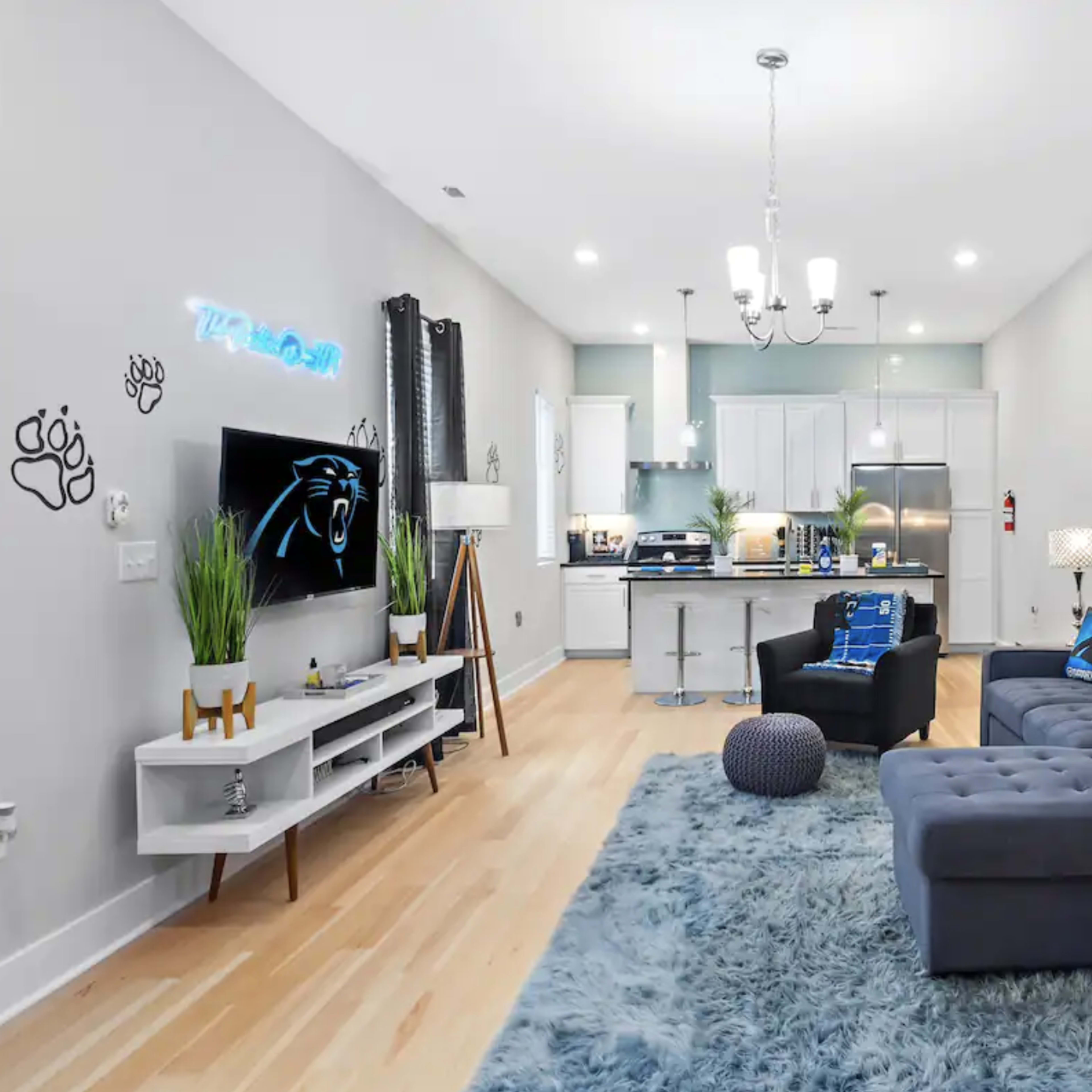A modern living room with a blue-themed decor, featuring a sectional sofa, a large area rug, and a mural on the wall, all adjacent to a kitchen with white cabinetry.