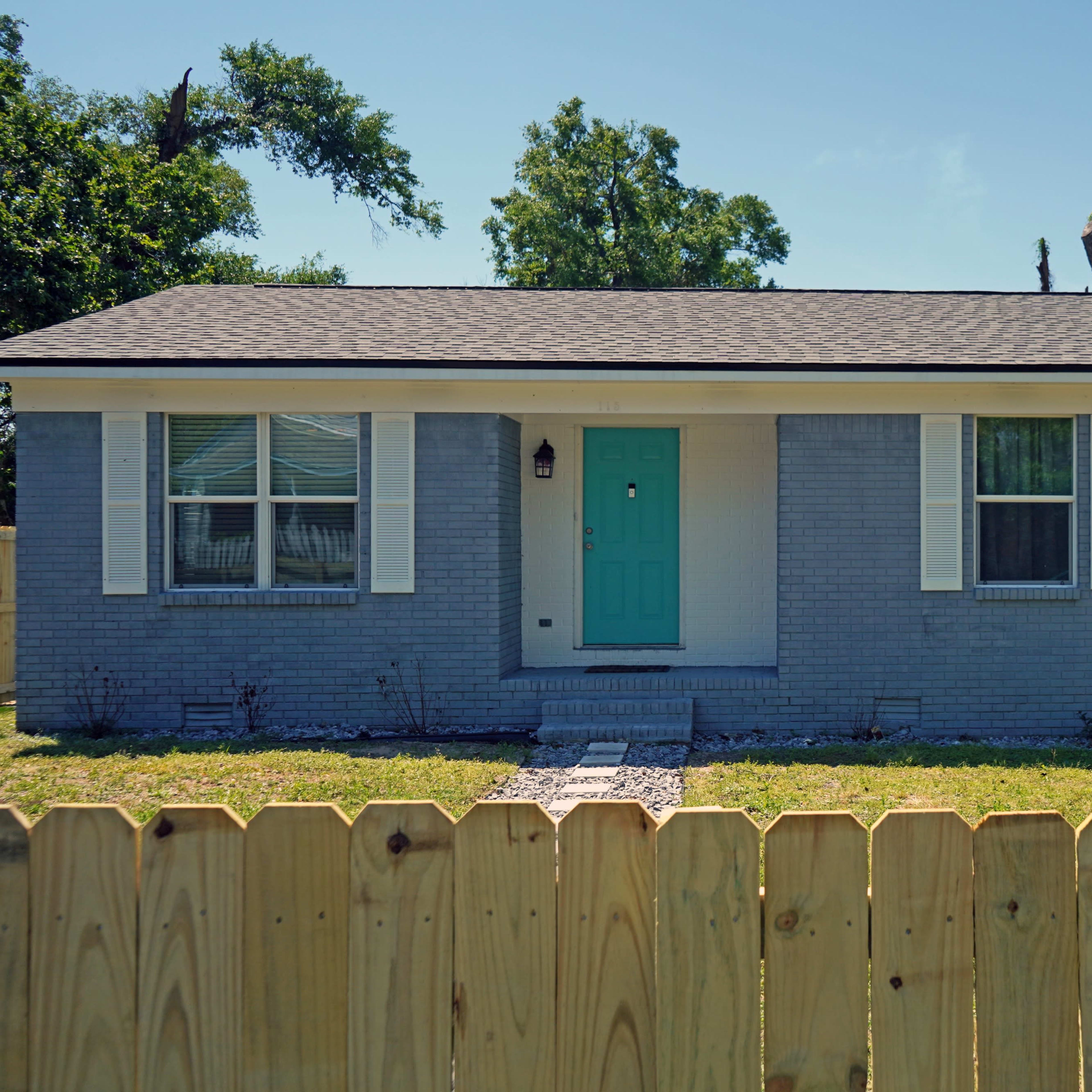 A single-story gray house with a turquoise front door and white shutters sits behind a wooden fence in a grassy yard.