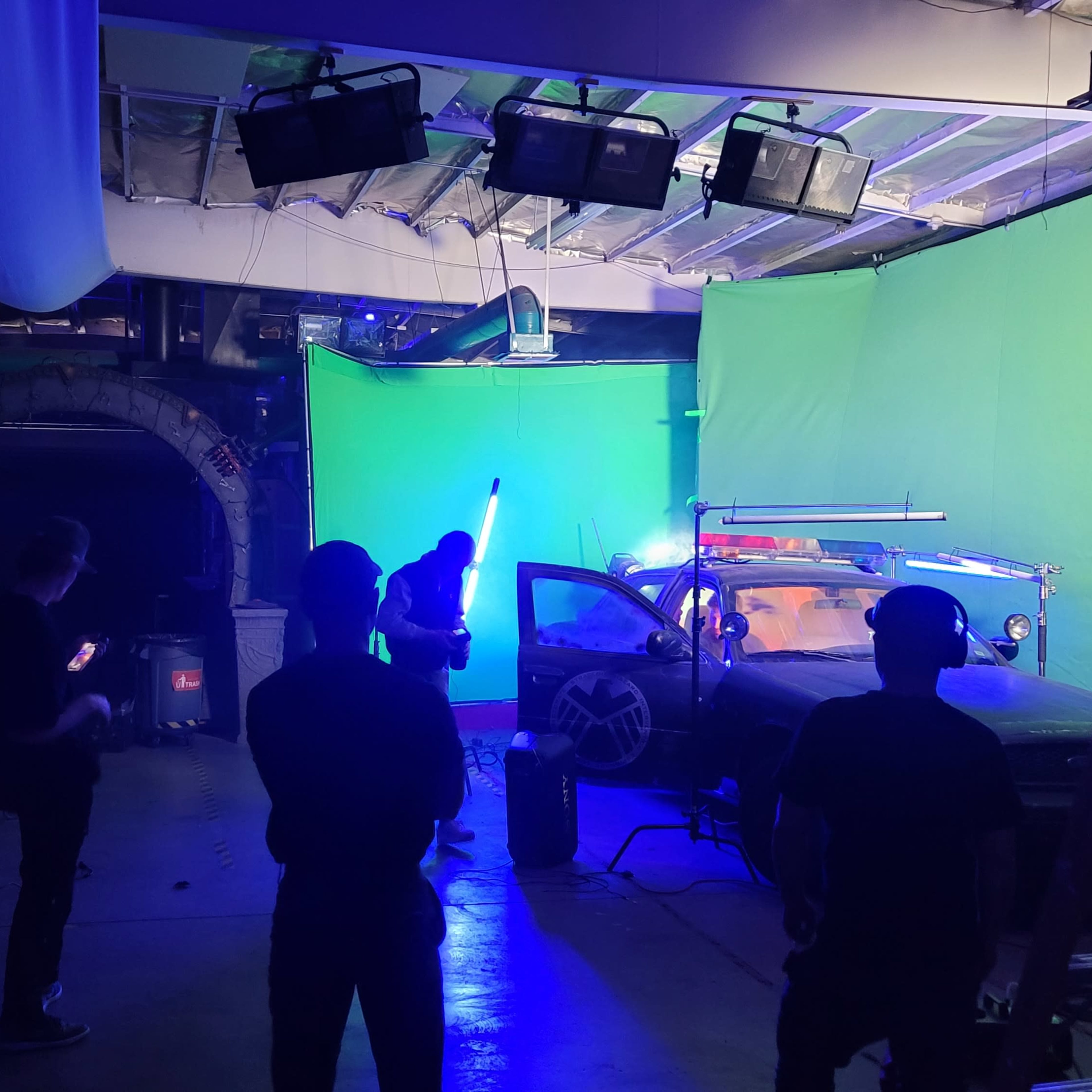 A film crew is working in a studio with a green screen, preparing equipment around a modified vehicle under artificial lighting.