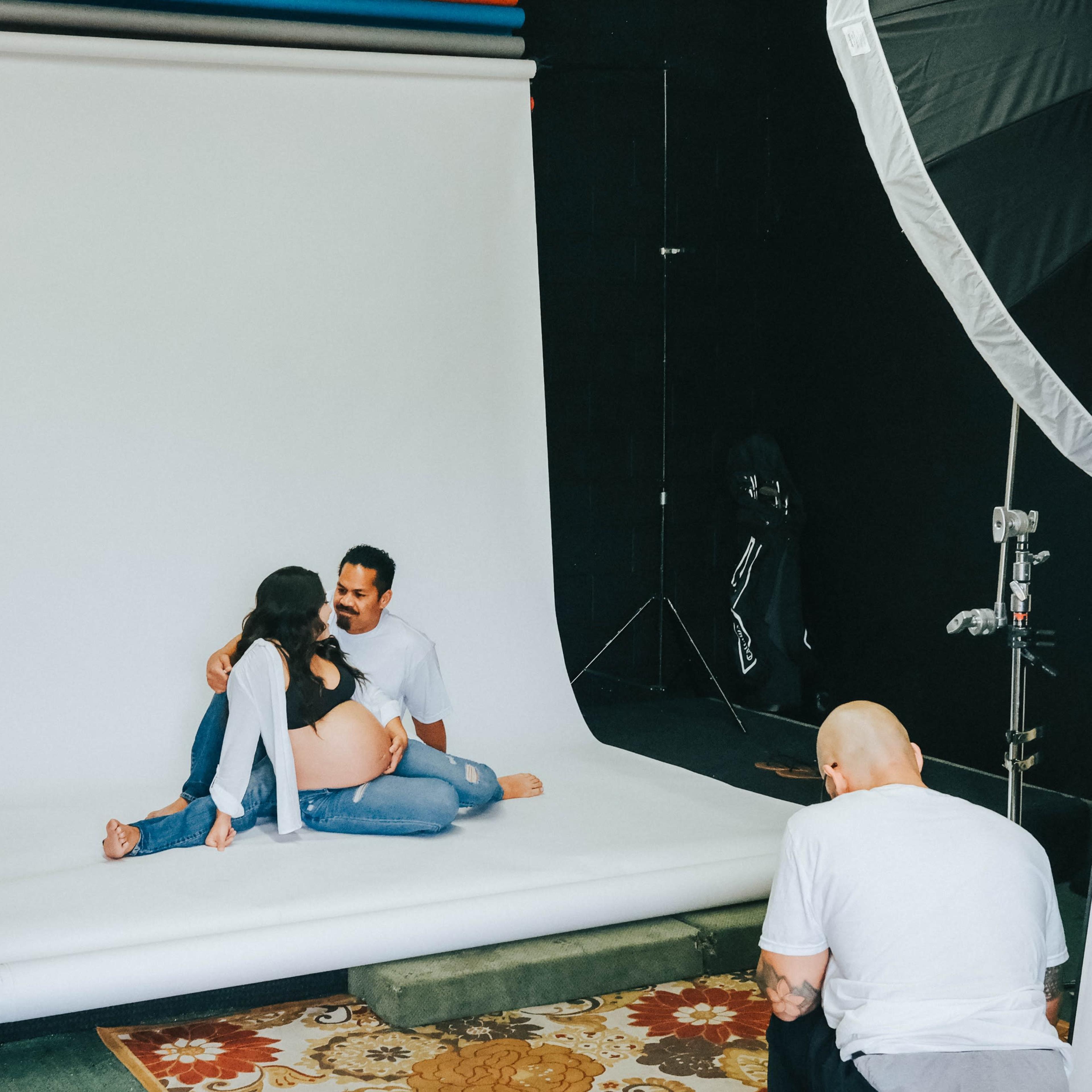 A couple poses for a photo shoot on a white backdrop while a photographer captures the scene from behind.