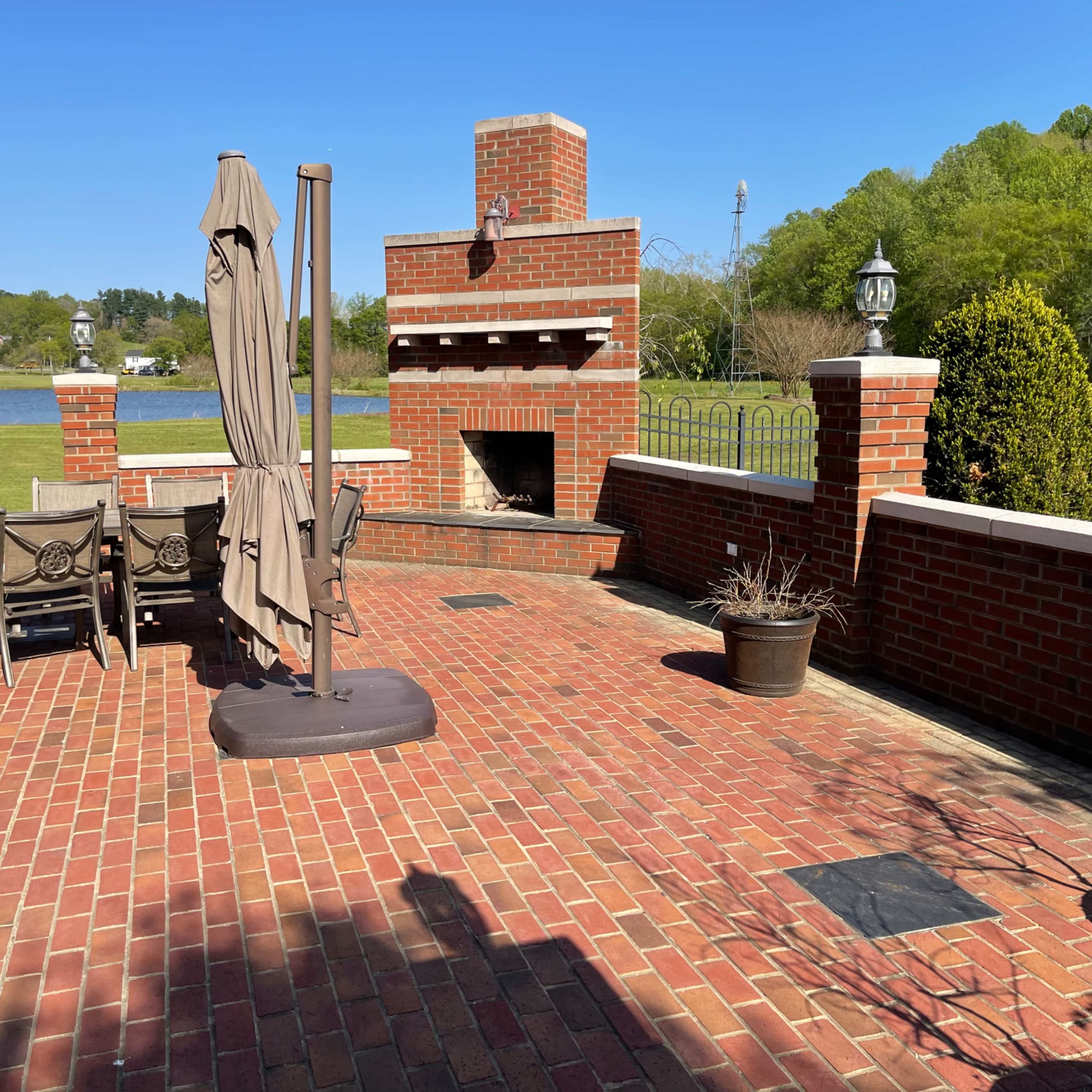 A brick patio features a fireplace, dining furniture, and a large umbrella, overlooking a grassy area and trees.