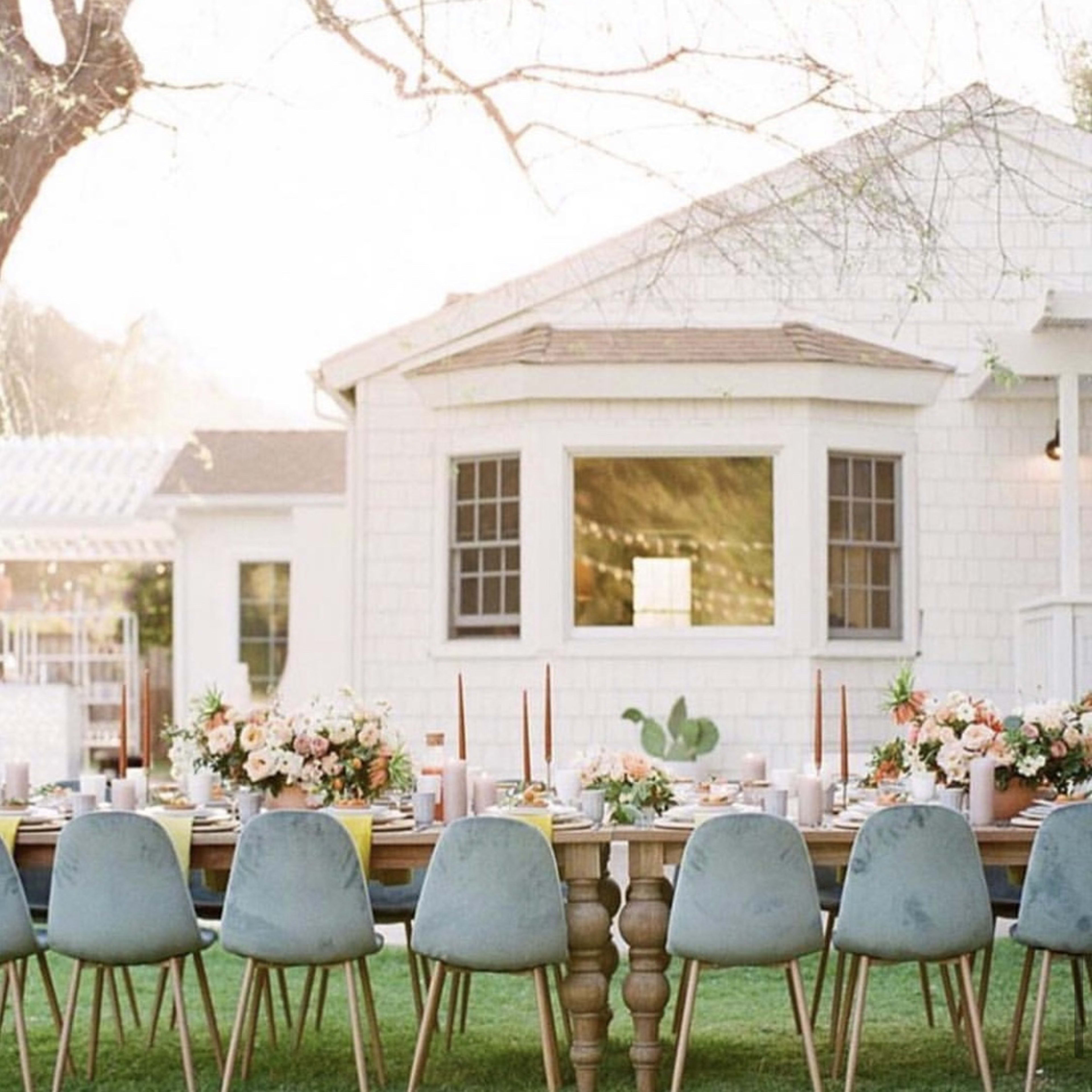 A long table is set with blue chairs and floral arrangements in a backyard near a white house.