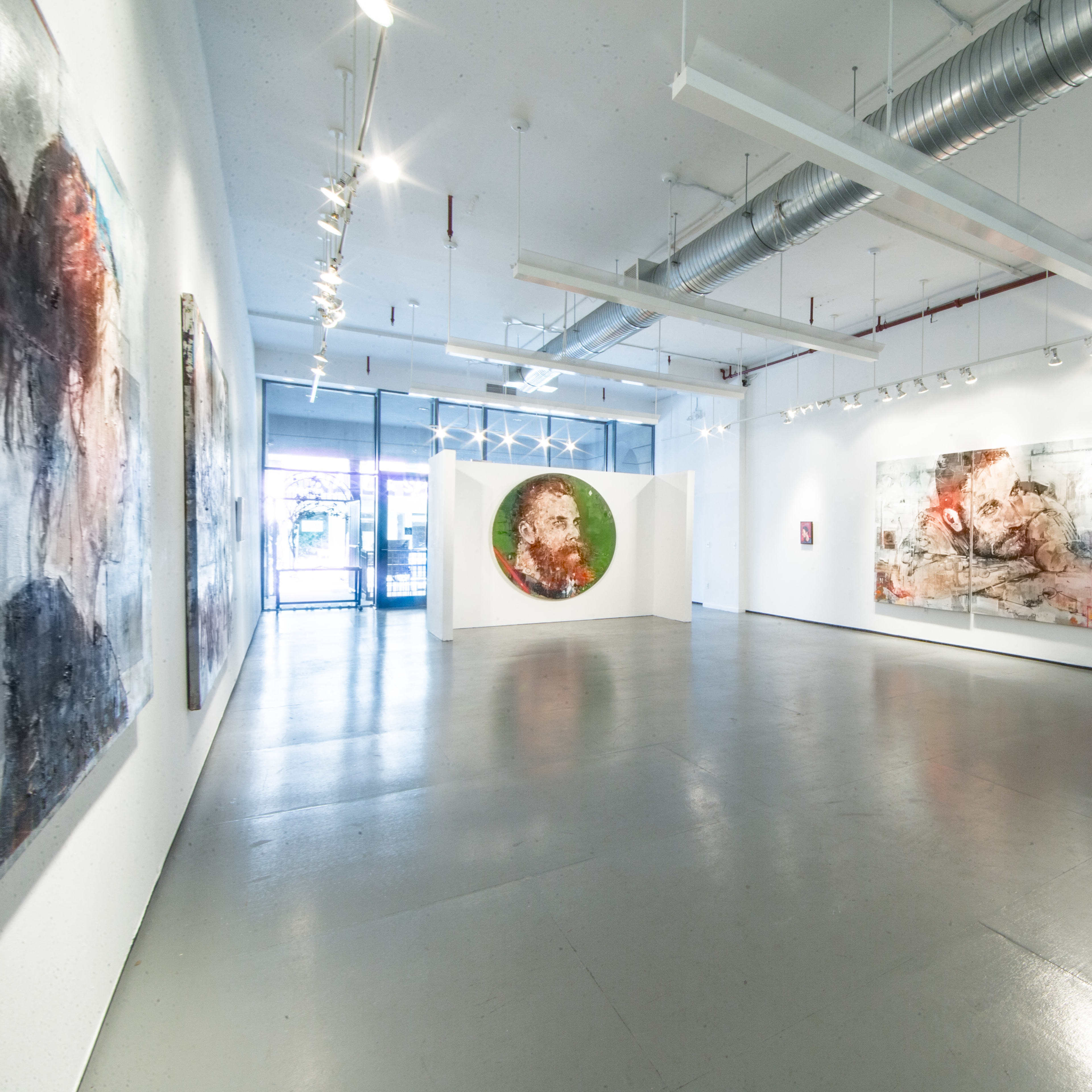 An art gallery features several large abstract paintings on white walls and a polished concrete floor, with natural light coming in from large windows.