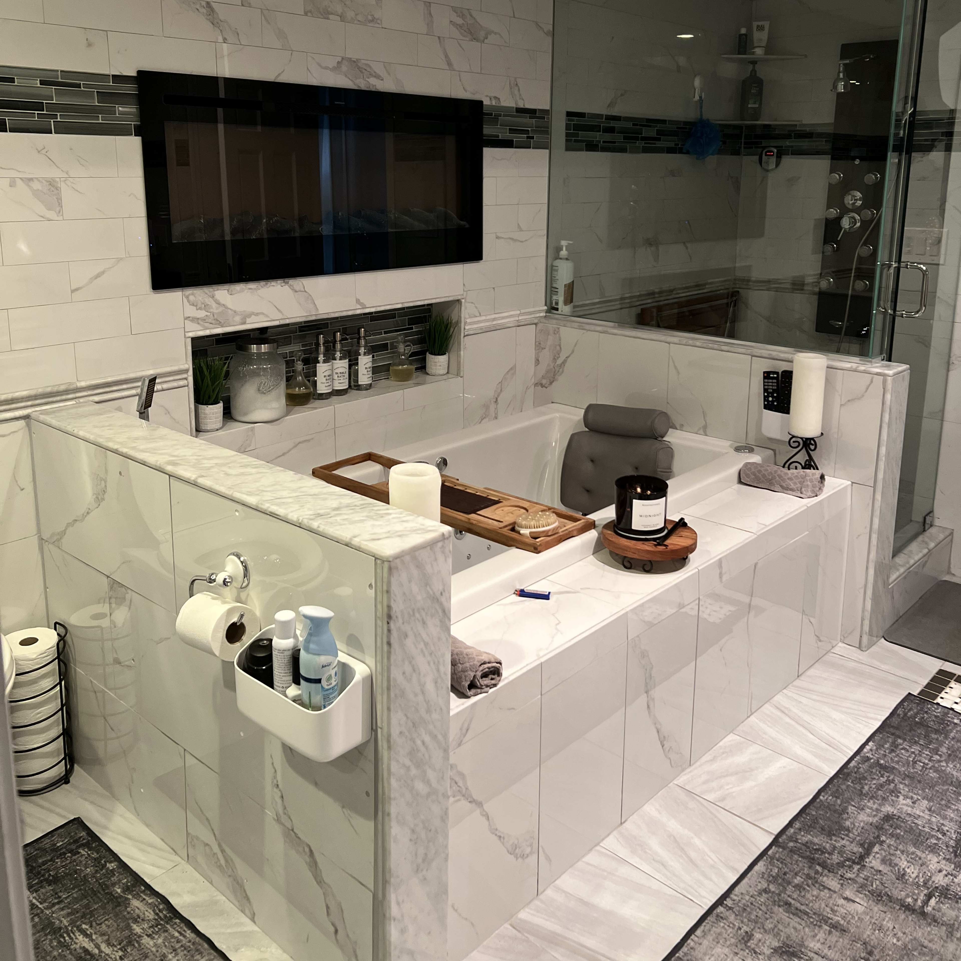 A modern bathroom featuring a large soaking tub with a wooden tray, a glass shower enclosure, and a tiled wall with decorative accents.