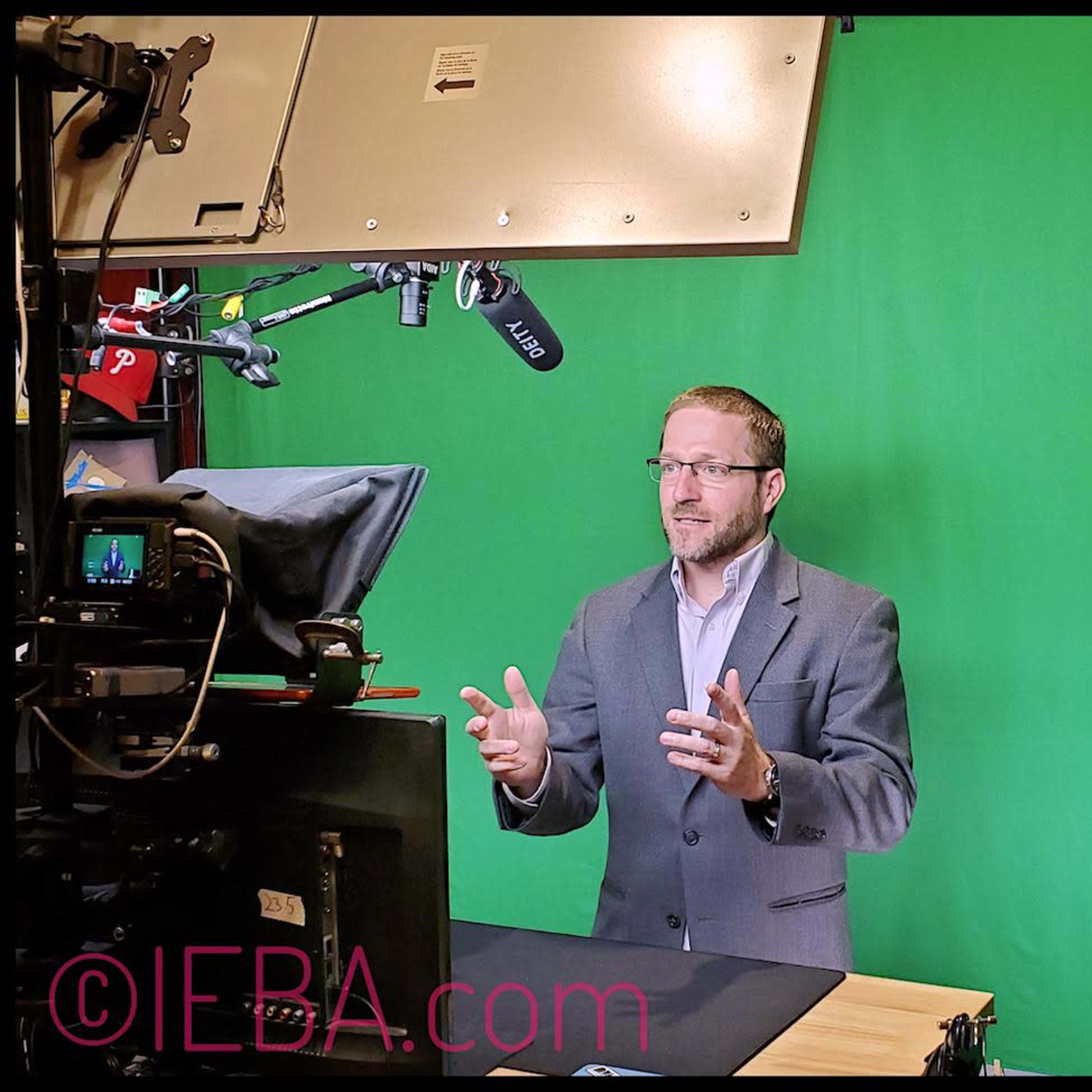 A man in a gray suit stands in front of a green screen while gesturing and speaking into a microphone.