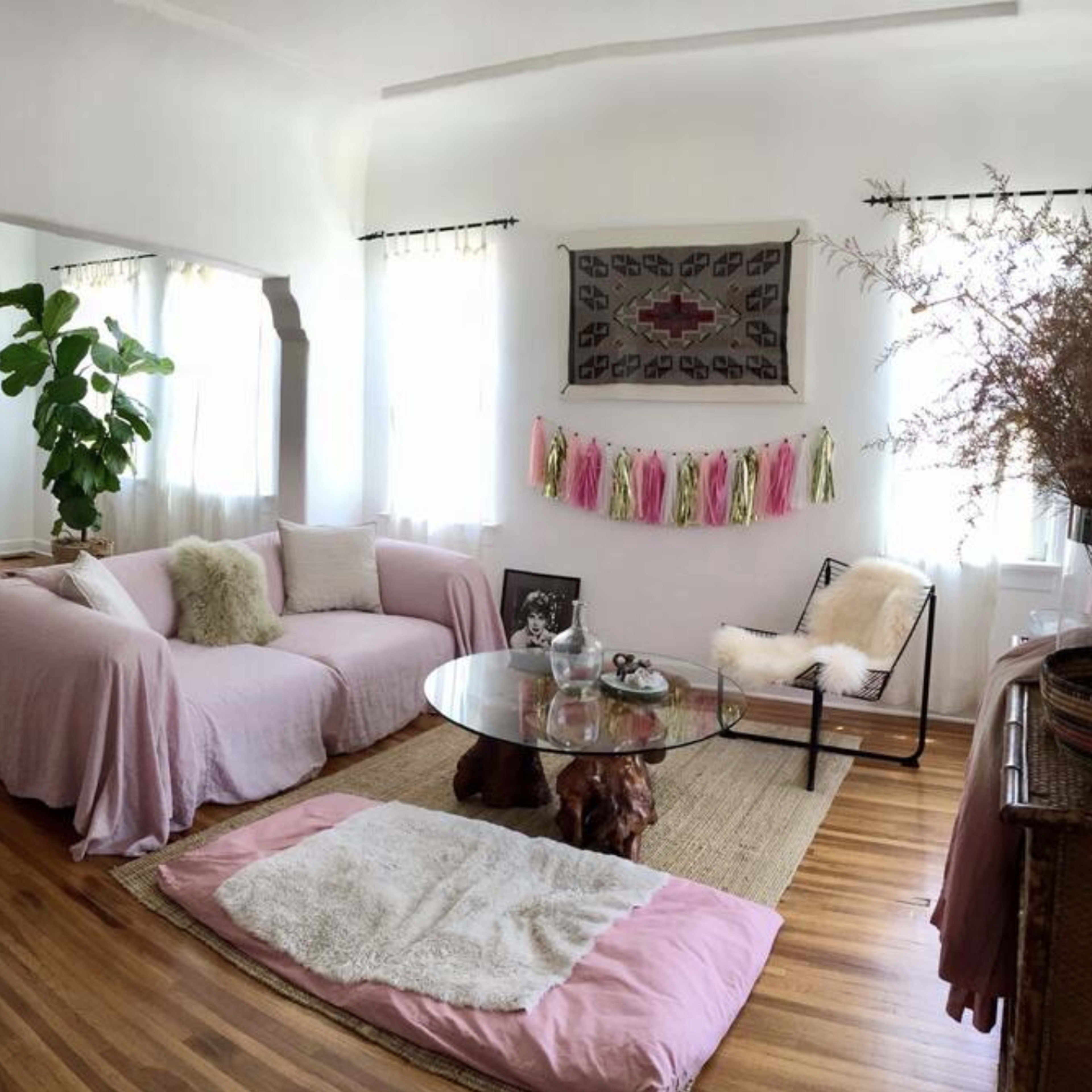 A living room with a pink sofa, wooden floor, a glass coffee table, and a dining area visible in the background.
