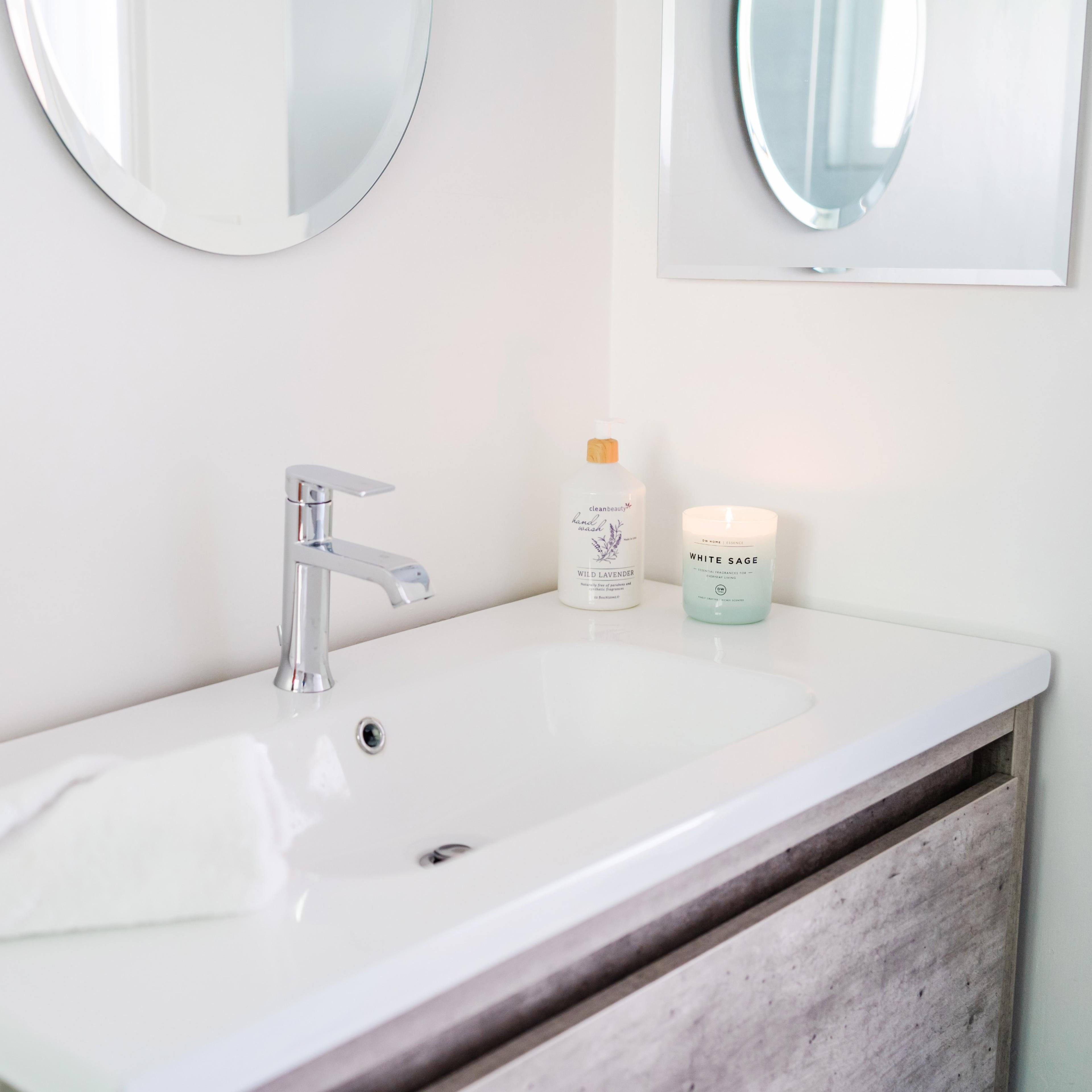 A modern bathroom vanity features a sink, a small potted cactus, a candle, a soap dispenser, and a neatly folded towel.