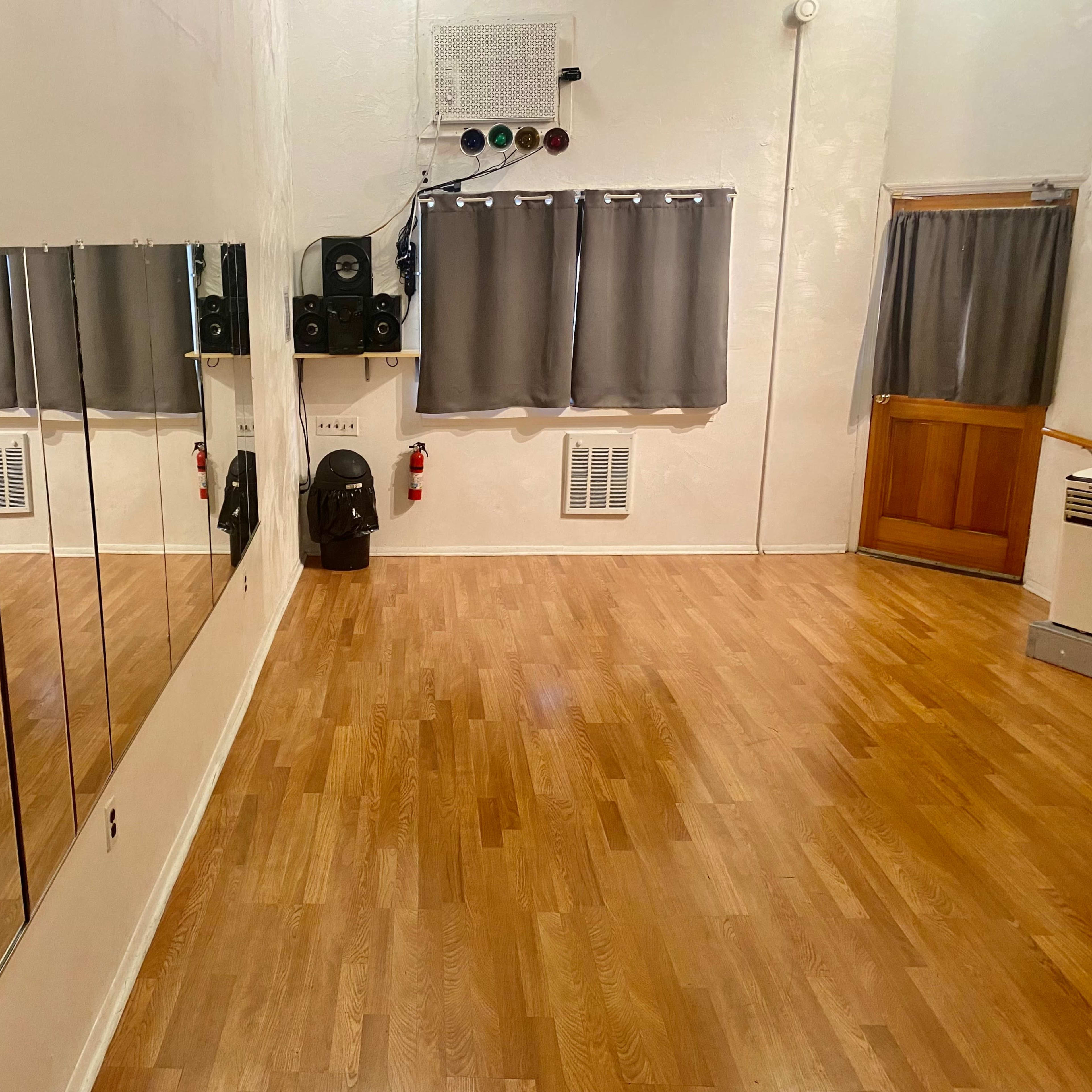 A dance studio with wooden flooring, mirrors on one wall, gray curtains, a door, and some audio equipment.