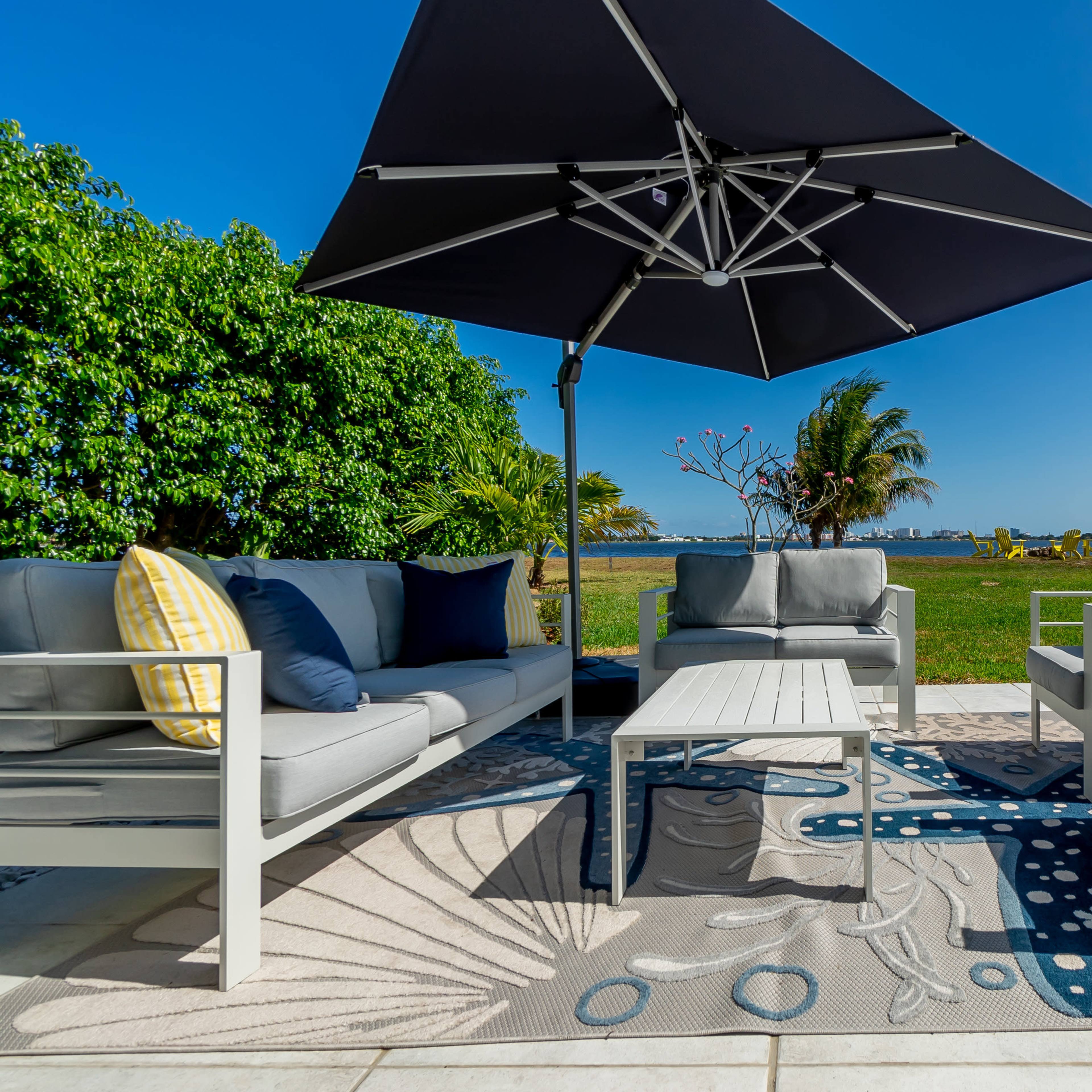A modern outdoor seating area features a large umbrella, gray sofas with colorful cushions, and a coffee table on a patterned rug, overlooking a lush green landscape.