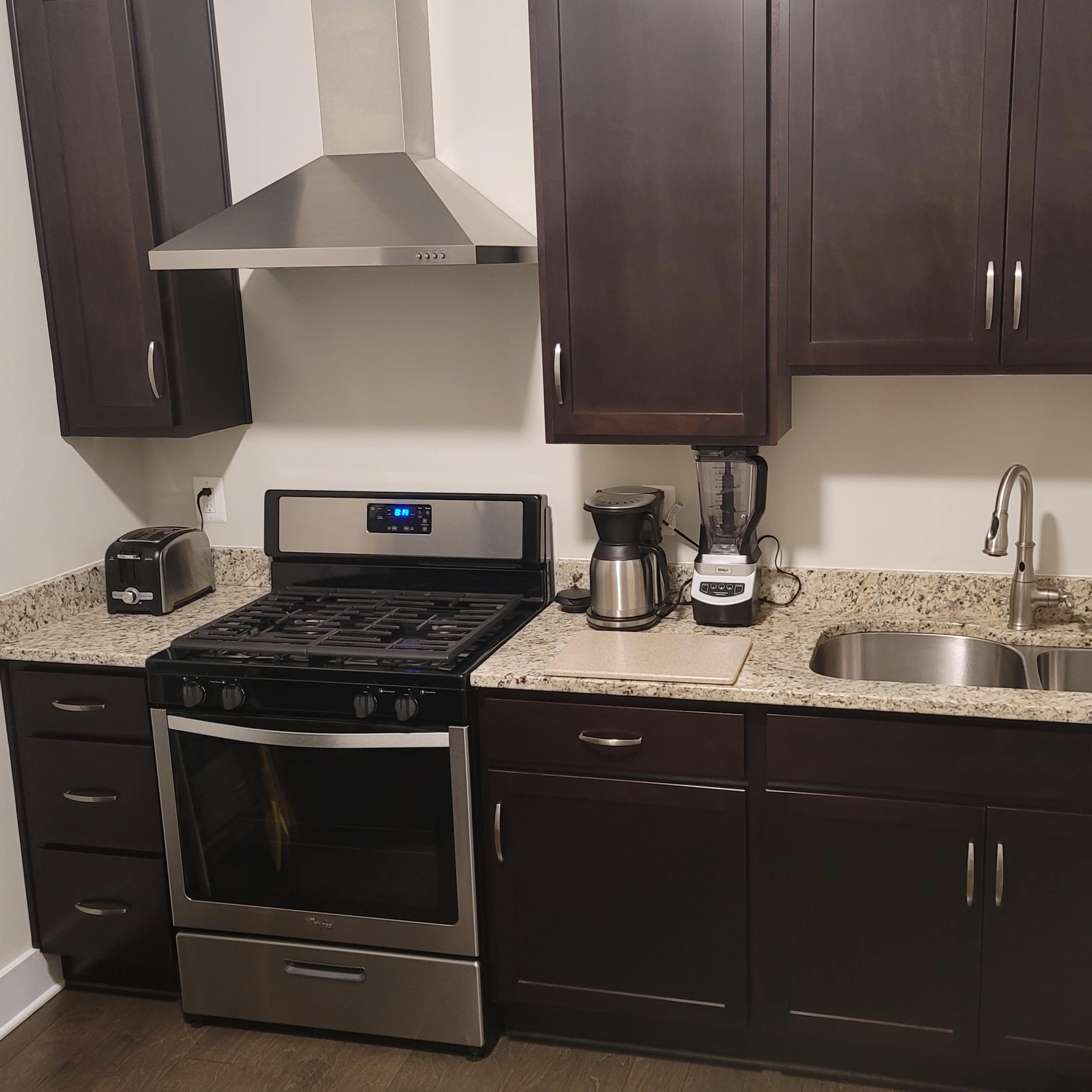 A compact kitchen features dark wooden cabinets, a granite countertop, a stainless steel stove, and a double sink with a pull-down faucet.