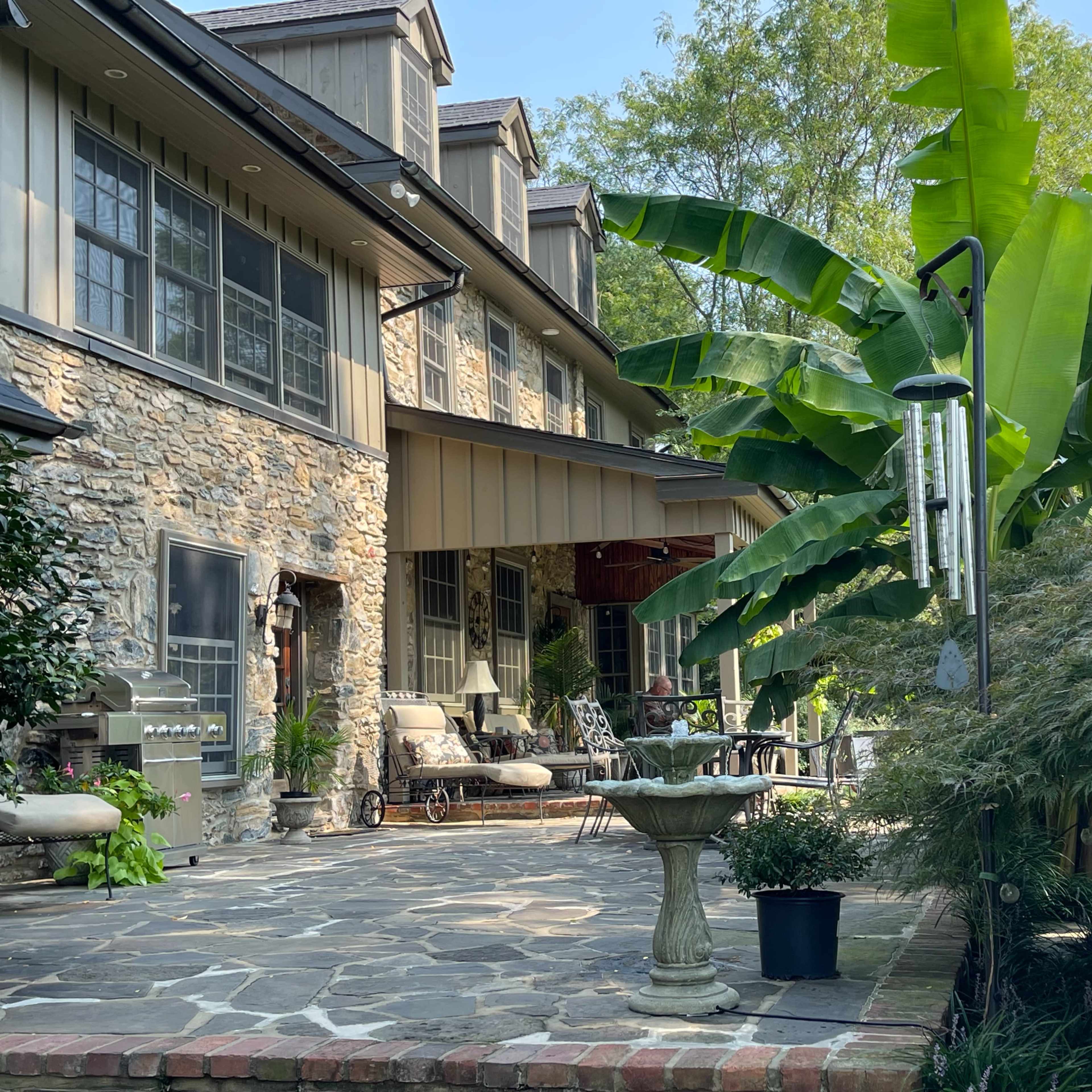 A stone patio with outdoor seating and a birdbath is situated beside large banana plants and wind chimes near a stone and wooden house.