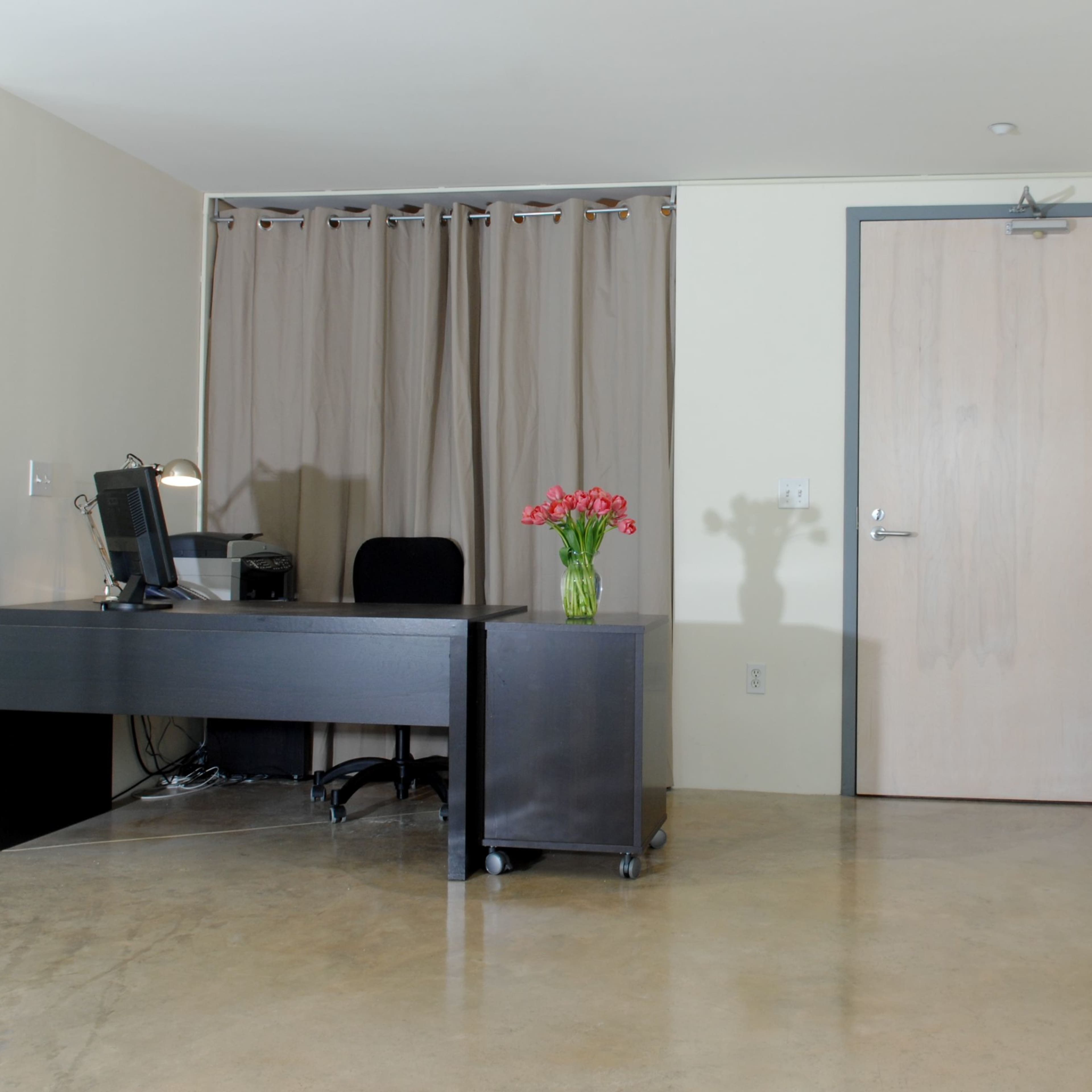 A minimalist office space with a desk, a computer, a chair, a vase of flowers, a closed door, and a large framed picture leaning against the wall.