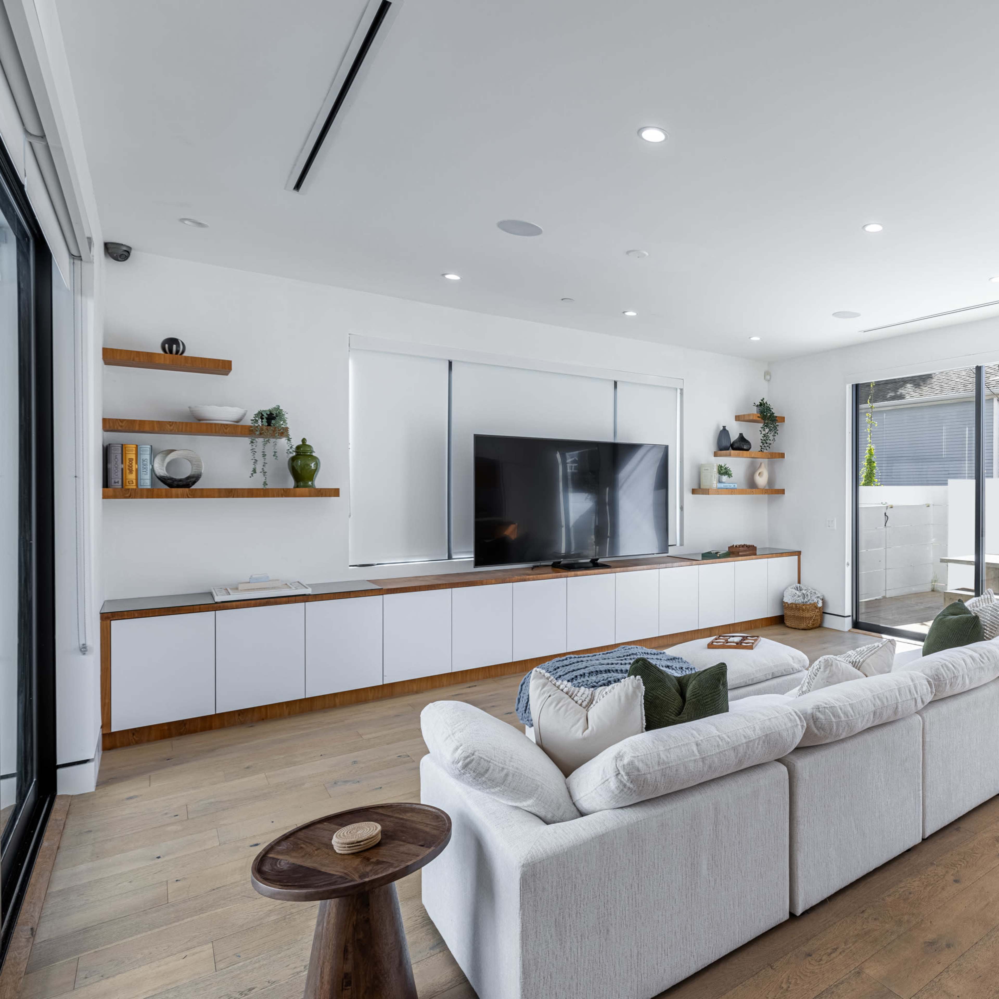A modern, bright living room with a large TV mounted on the wall, a white sectional sofa, and sliding glass doors leading to an outdoor area.