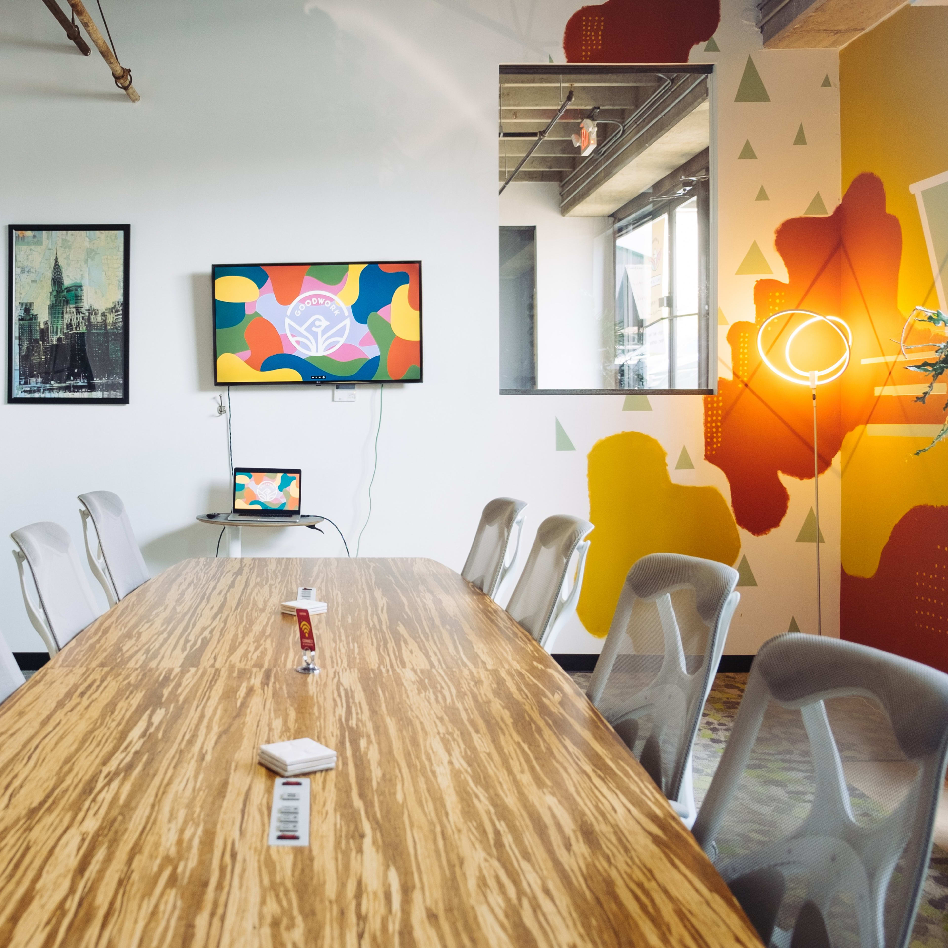 A modern conference room features a long wooden table, several ergonomic chairs, a wall-mounted TV displaying colorful artwork, and vibrant wall decorations.