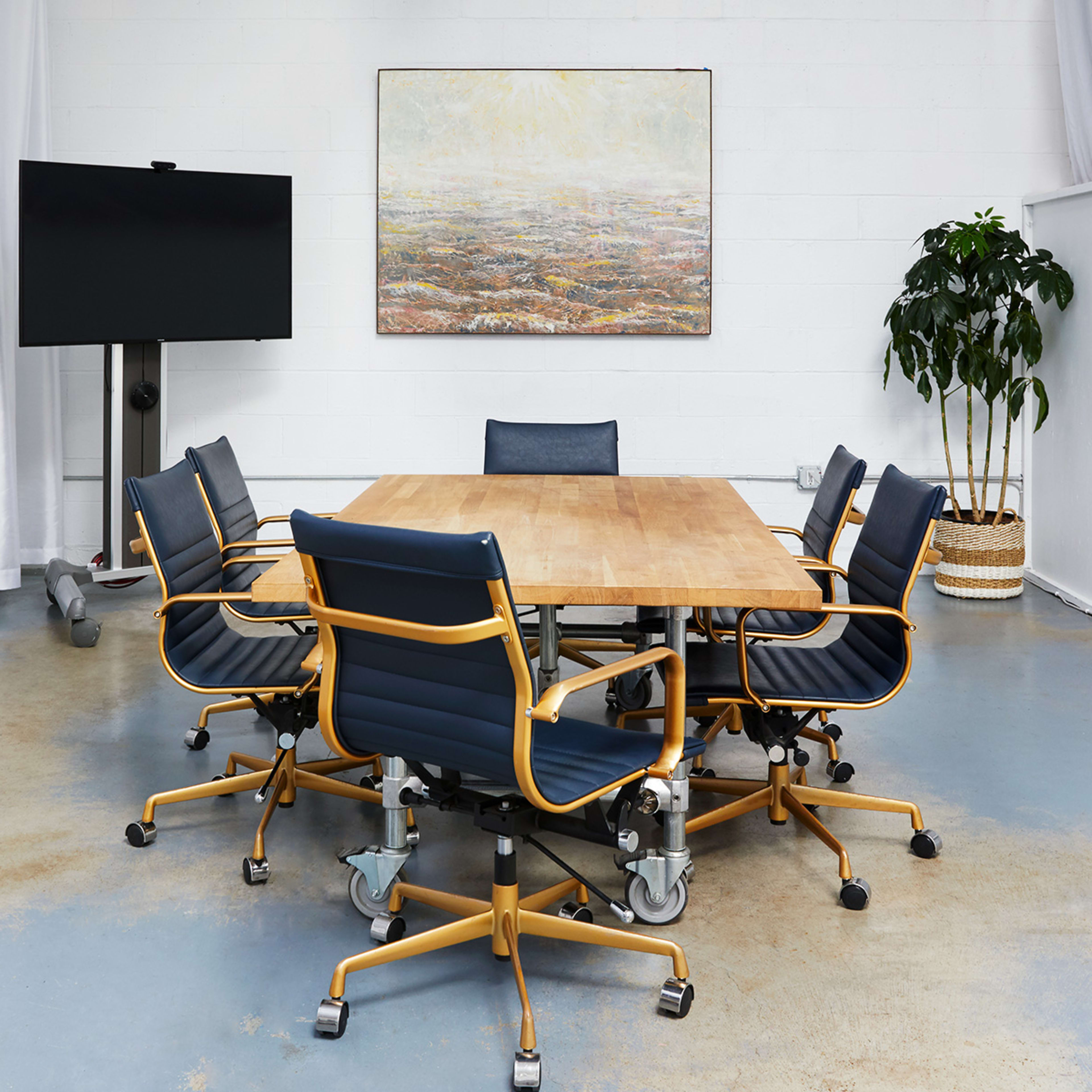 A modern conference room features a large wooden table surrounded by ergonomic chairs, with a television and a potted plant in the background.
