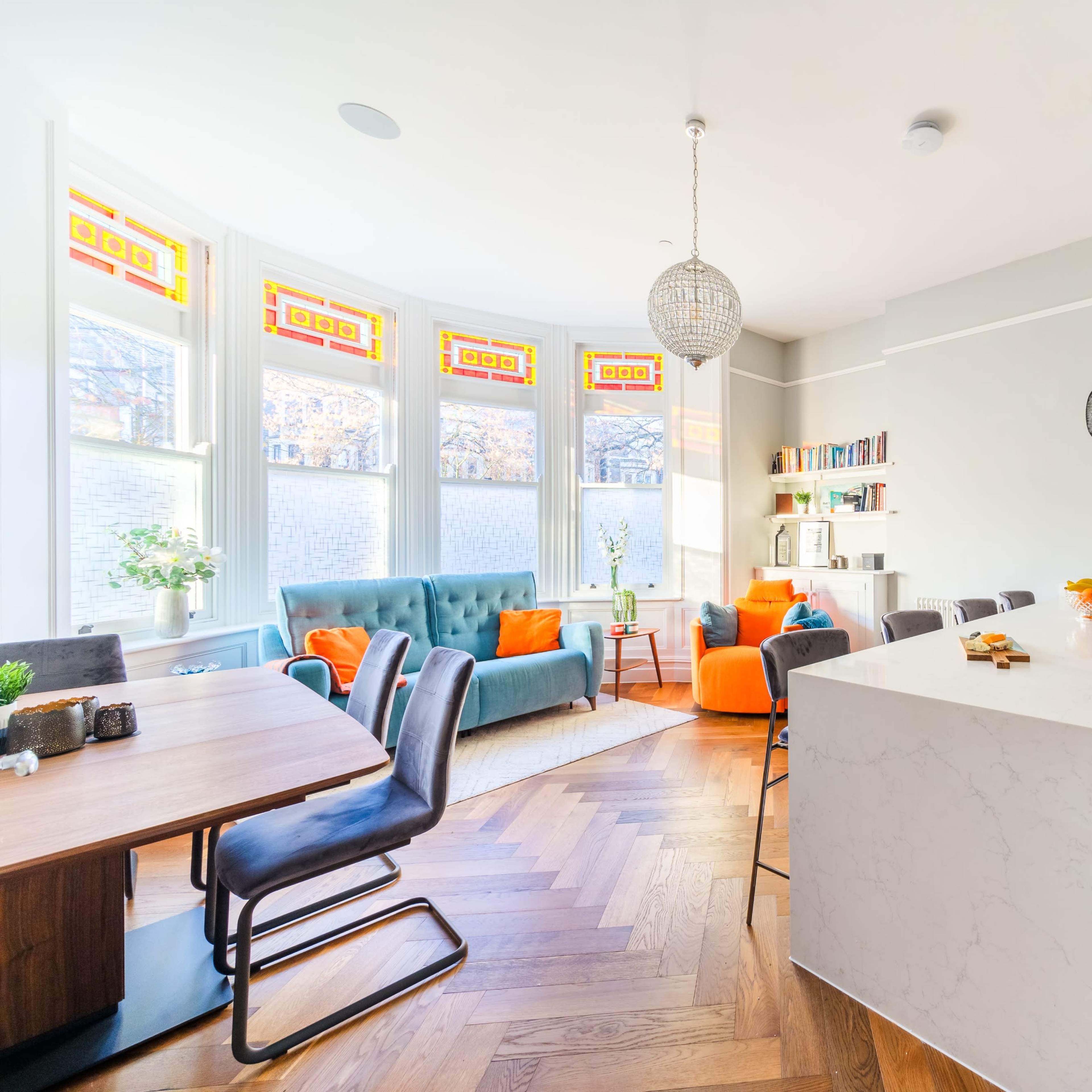 A bright open-plan kitchen and living area featuring a marble island, a wooden dining table, and stylish furniture in a room with large windows.