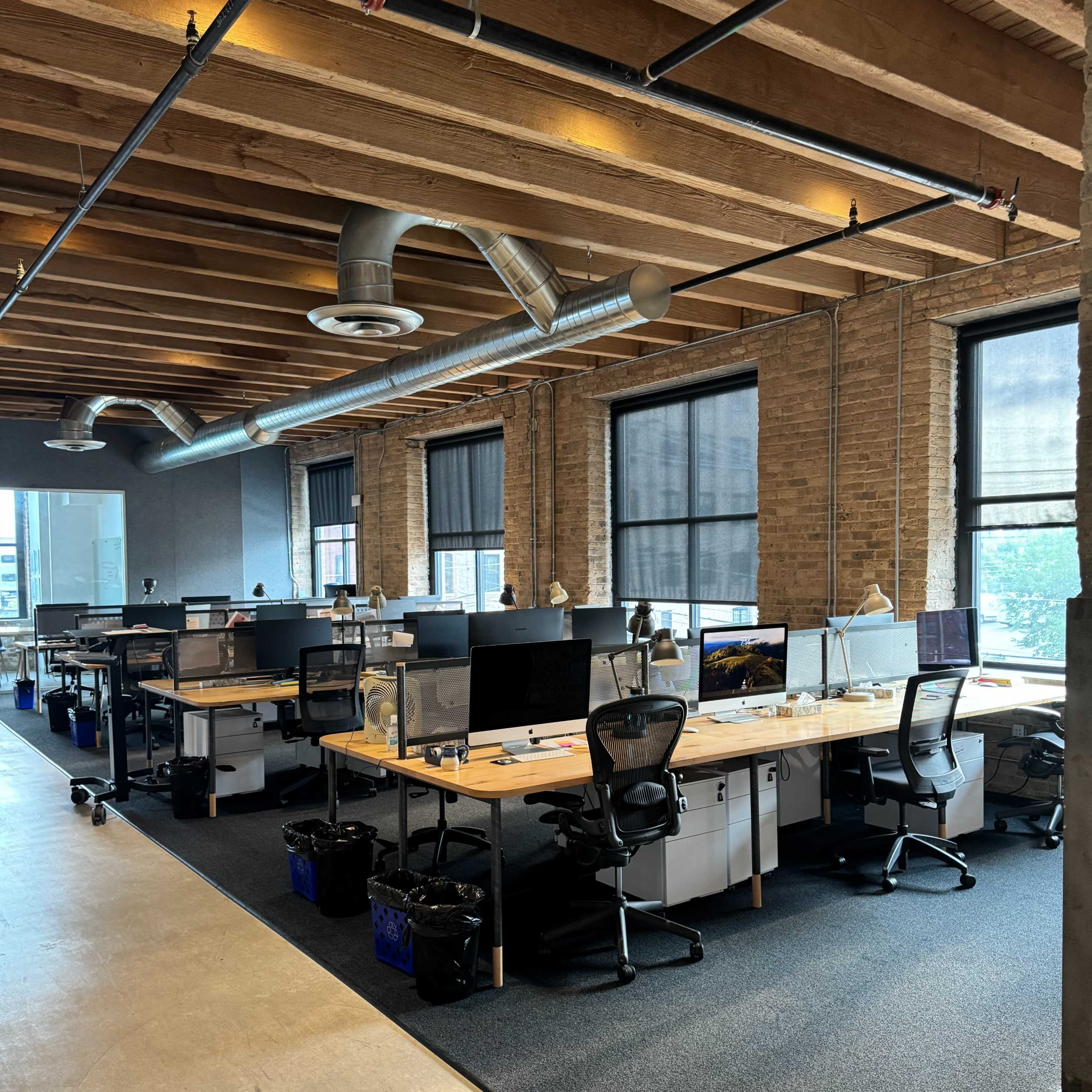 A modern office space with multiple desks, computers, and a carpeted floor under exposed wooden beams.