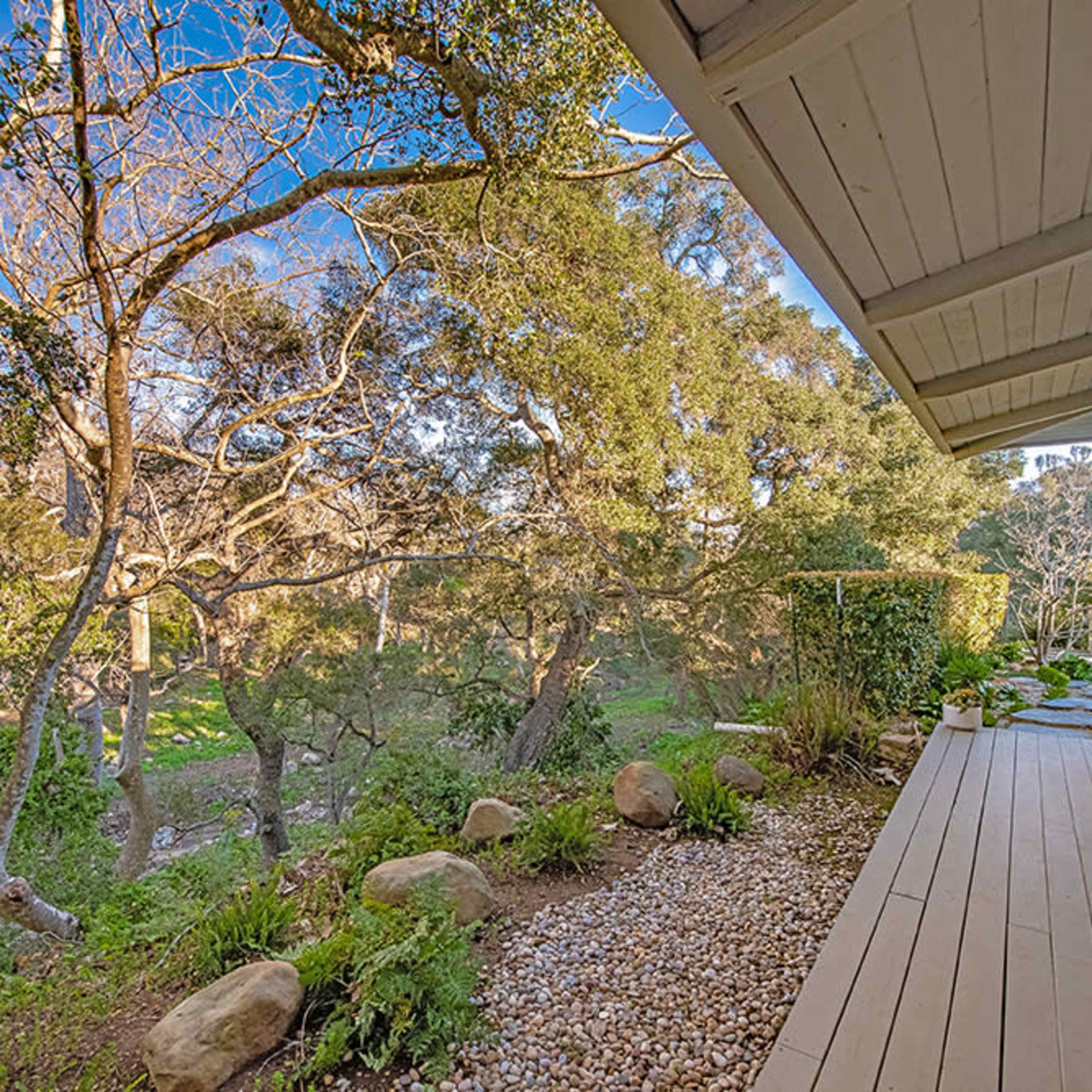 A wooden deck is surrounded by trees and greenery, with a path of pebbles and rocks leading into the landscape.
