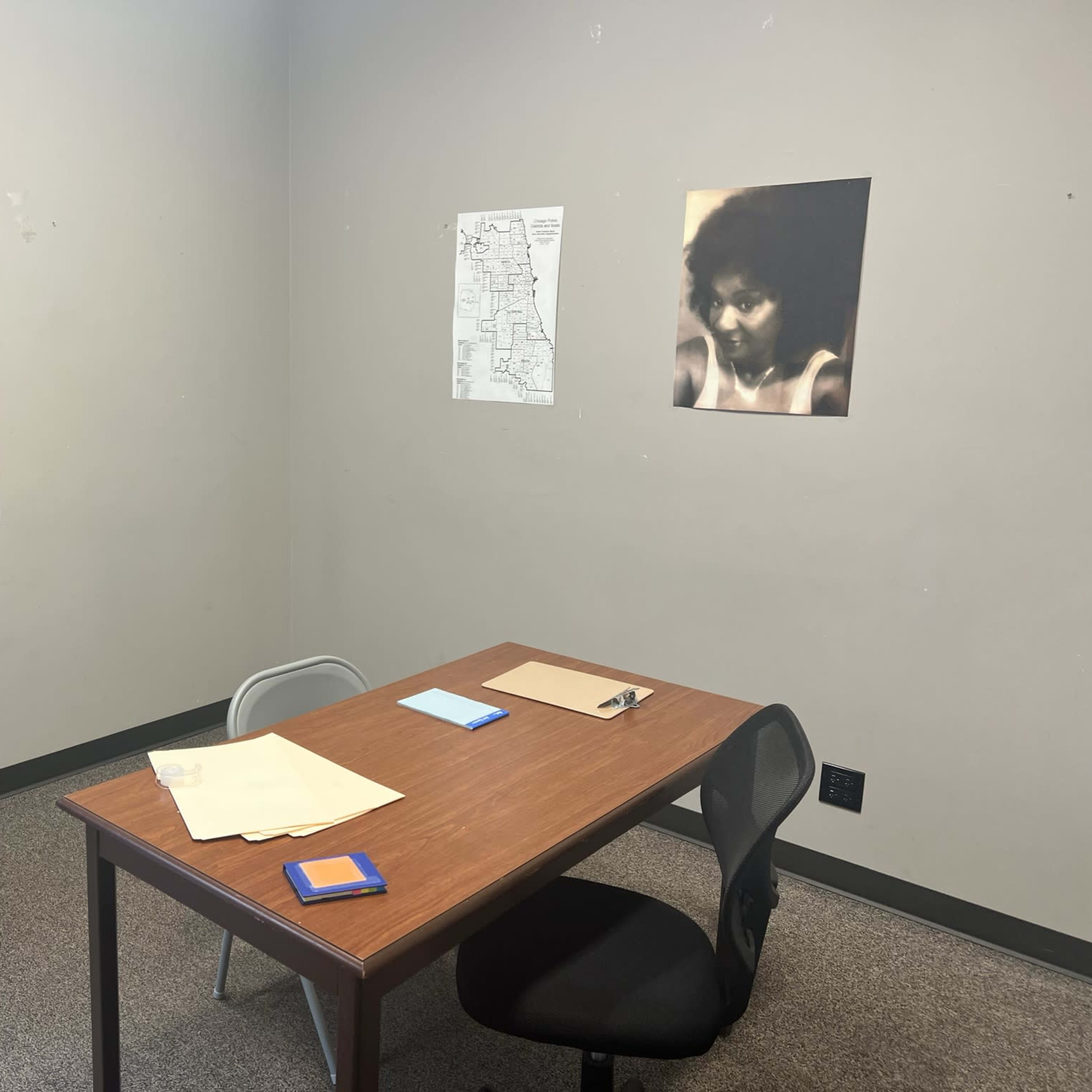 A plain room with a table and chair, two folders and a notepad on the table, a wall with a map and a photo of a person, and a window.