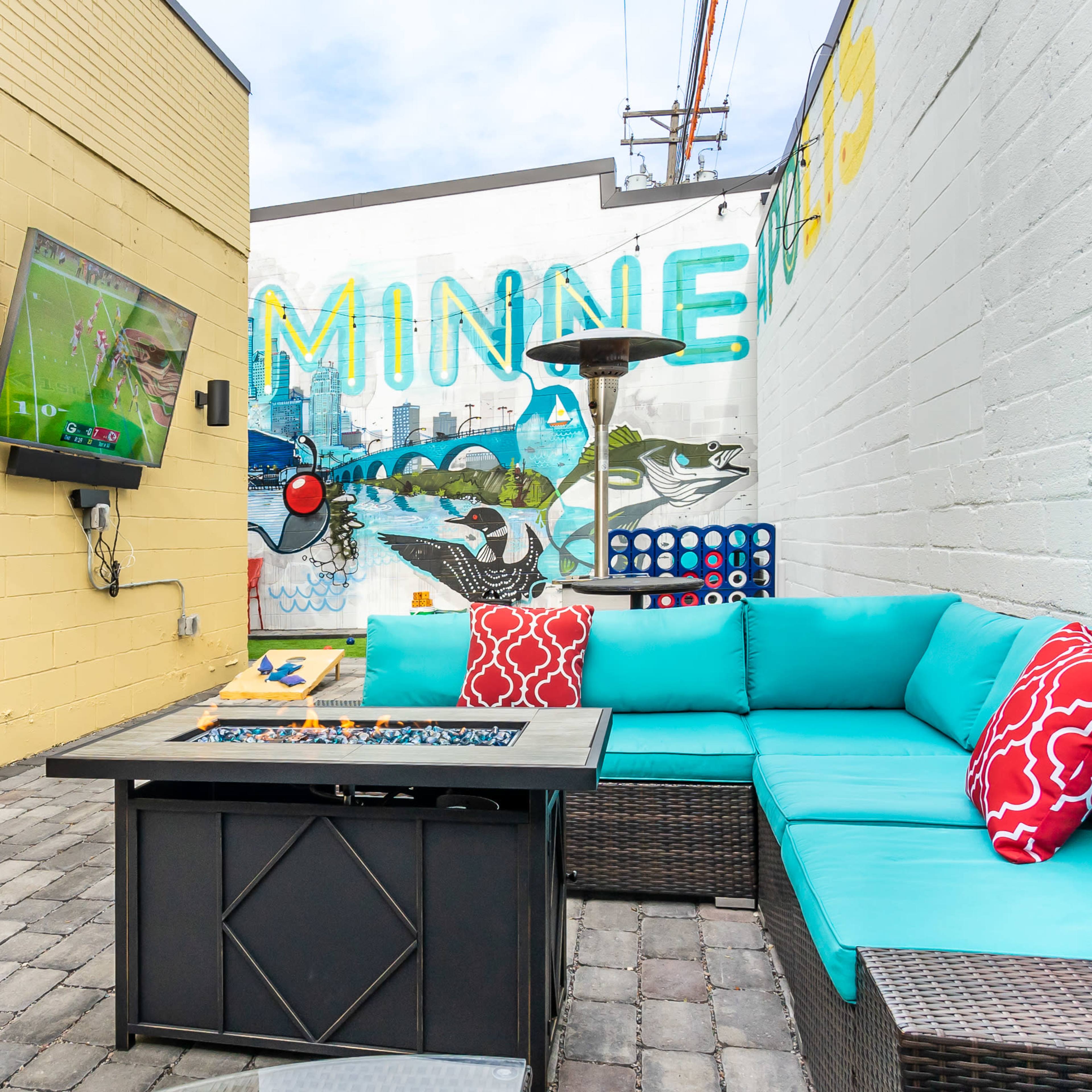 A patio area features a sectional sofa with red and turquoise cushions, a fire pit table, and a wall-mounted TV displaying a sports game, set against a colorful mural.