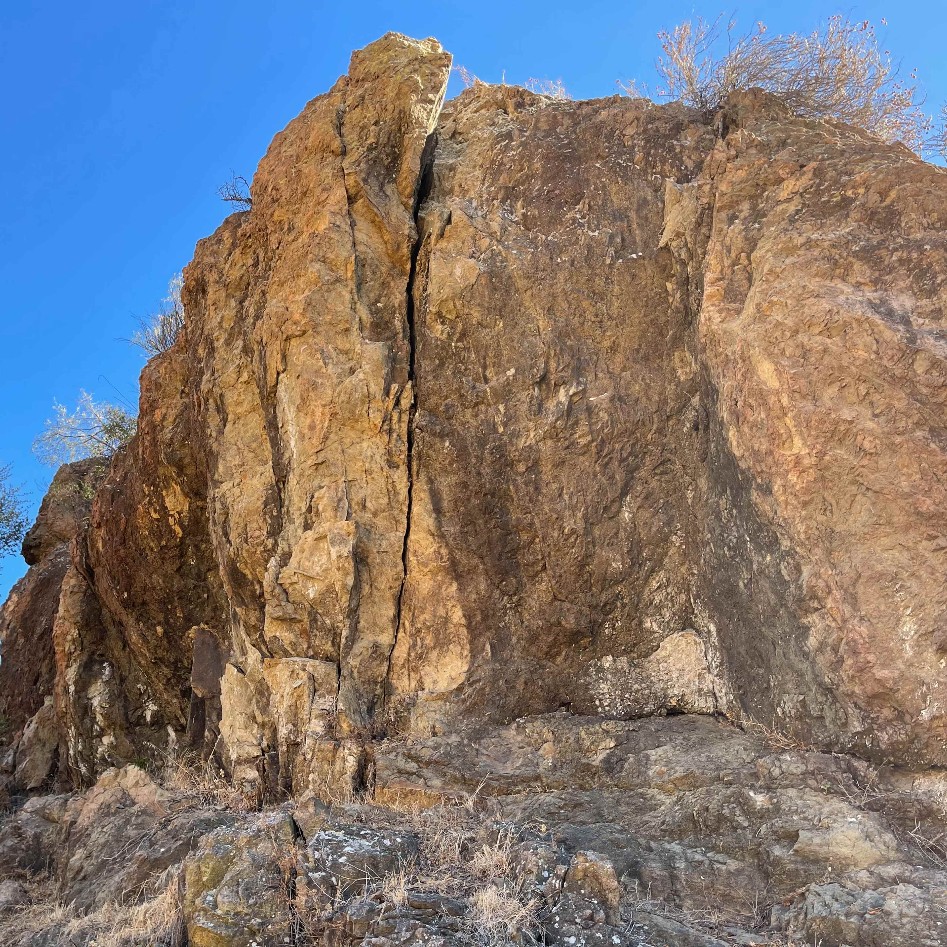 A large rock formation with a visible crack stands against a clear blue sky, surrounded by sparse vegetation.