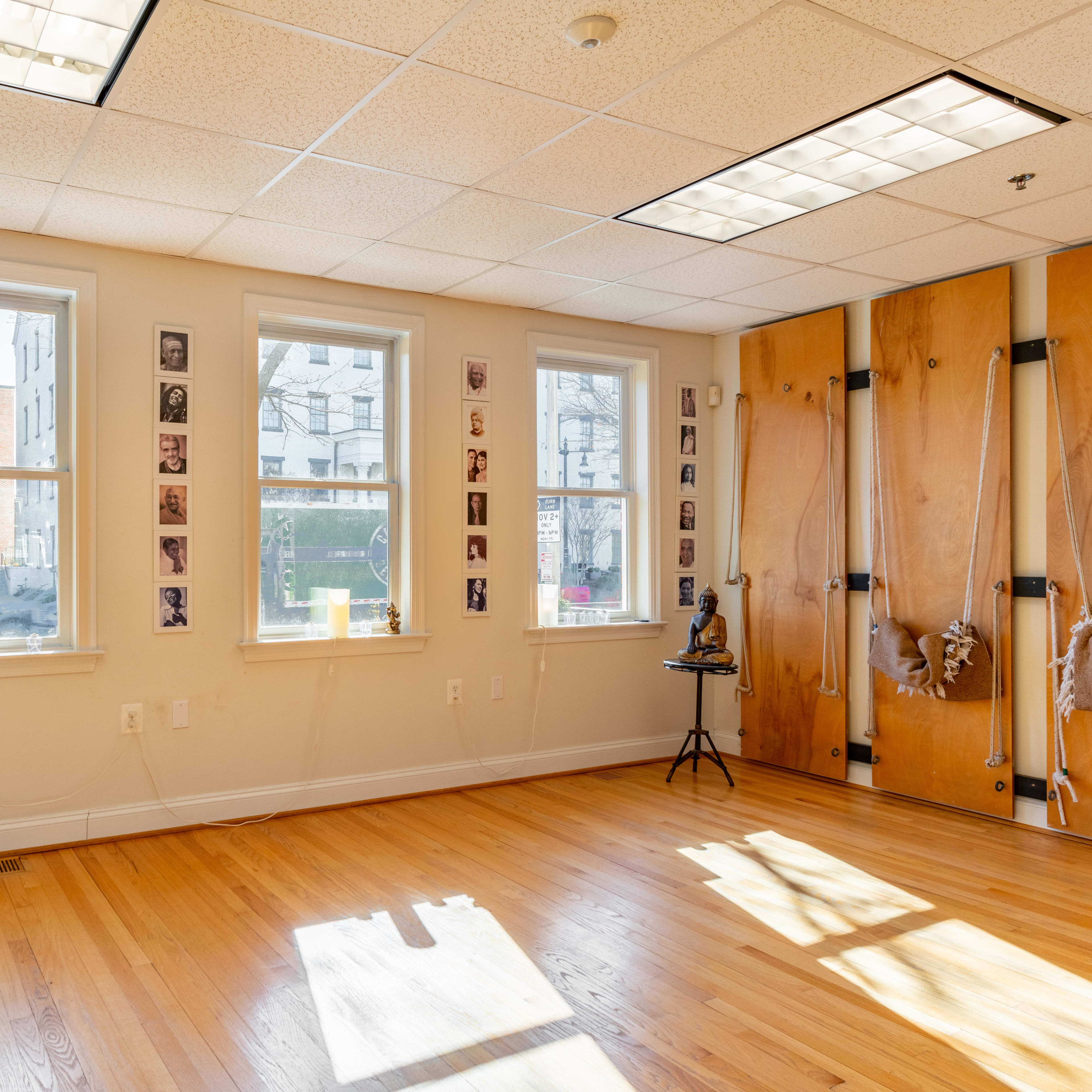 A spacious room with wooden flooring, featuring three windows, wall-mounted photographs, and a wooden partition on one side.