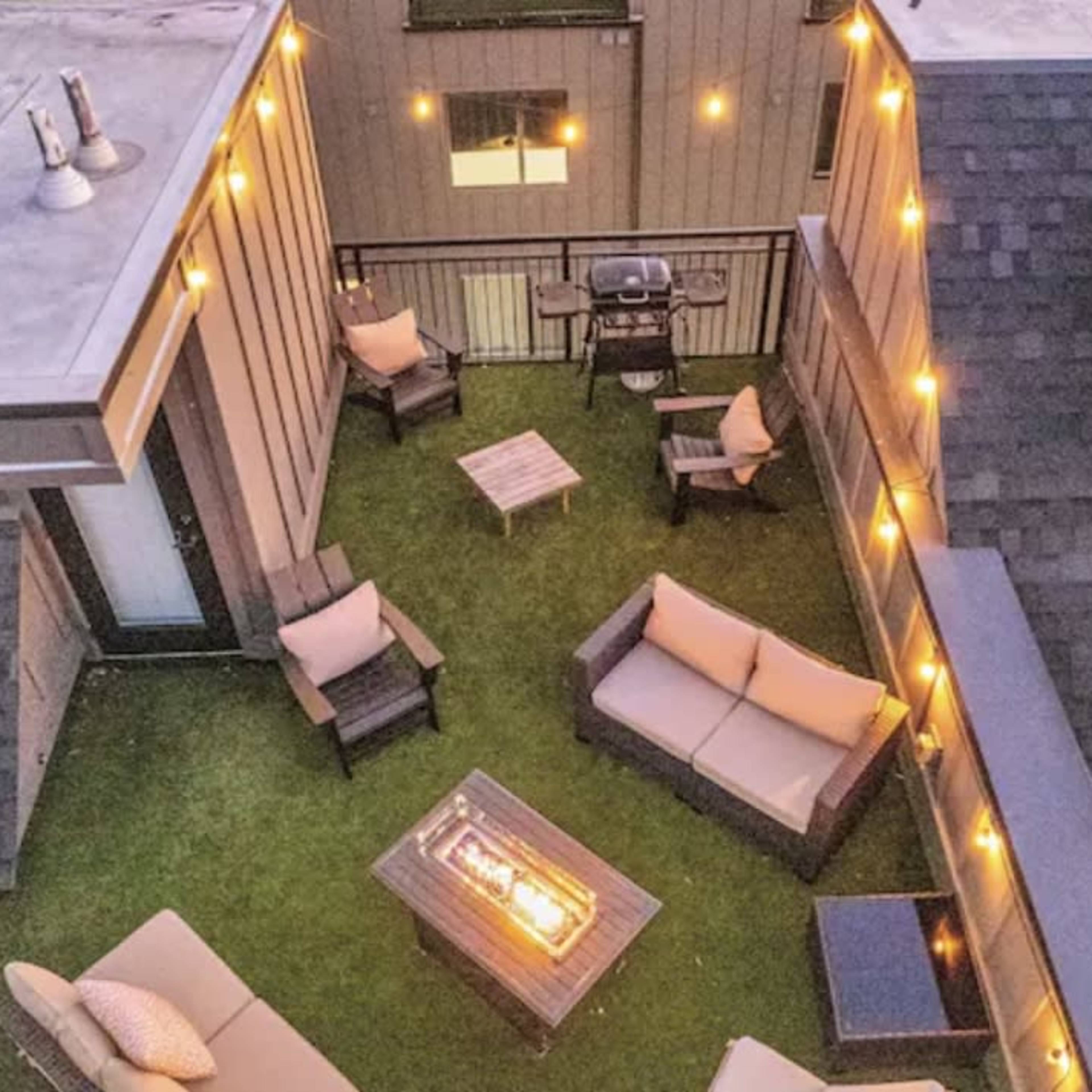 A rooftop terrace with outdoor furniture, string lights, and a grill, surrounded by buildings.