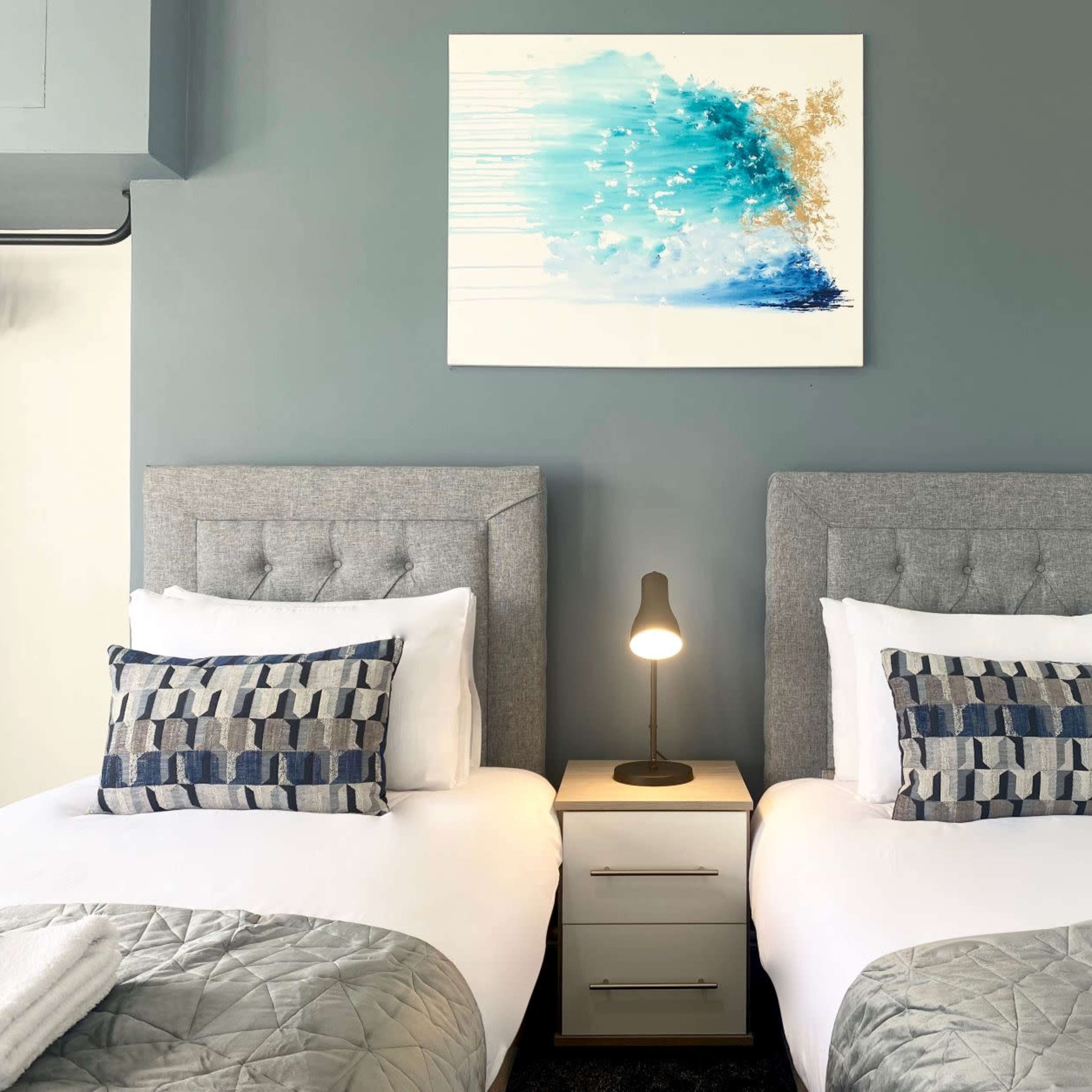 A bedroom with two twin beds, each with gray upholstered headboards, a bedside table with a lamp, and a piece of abstract art above the beds against a gray wall.
