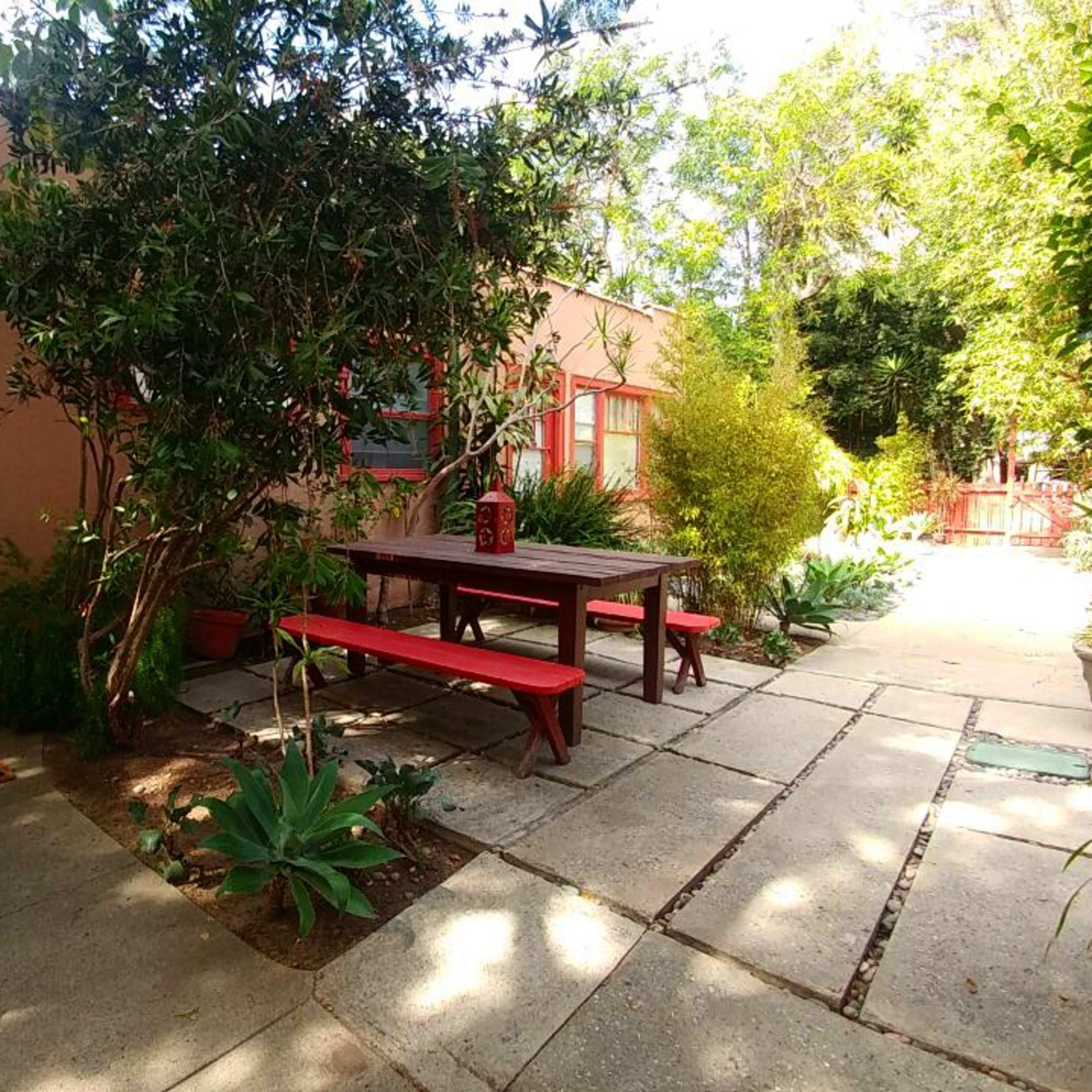 A patio area with a red picnic table surrounded by greenery and potted plants, leading to a building with a red door.