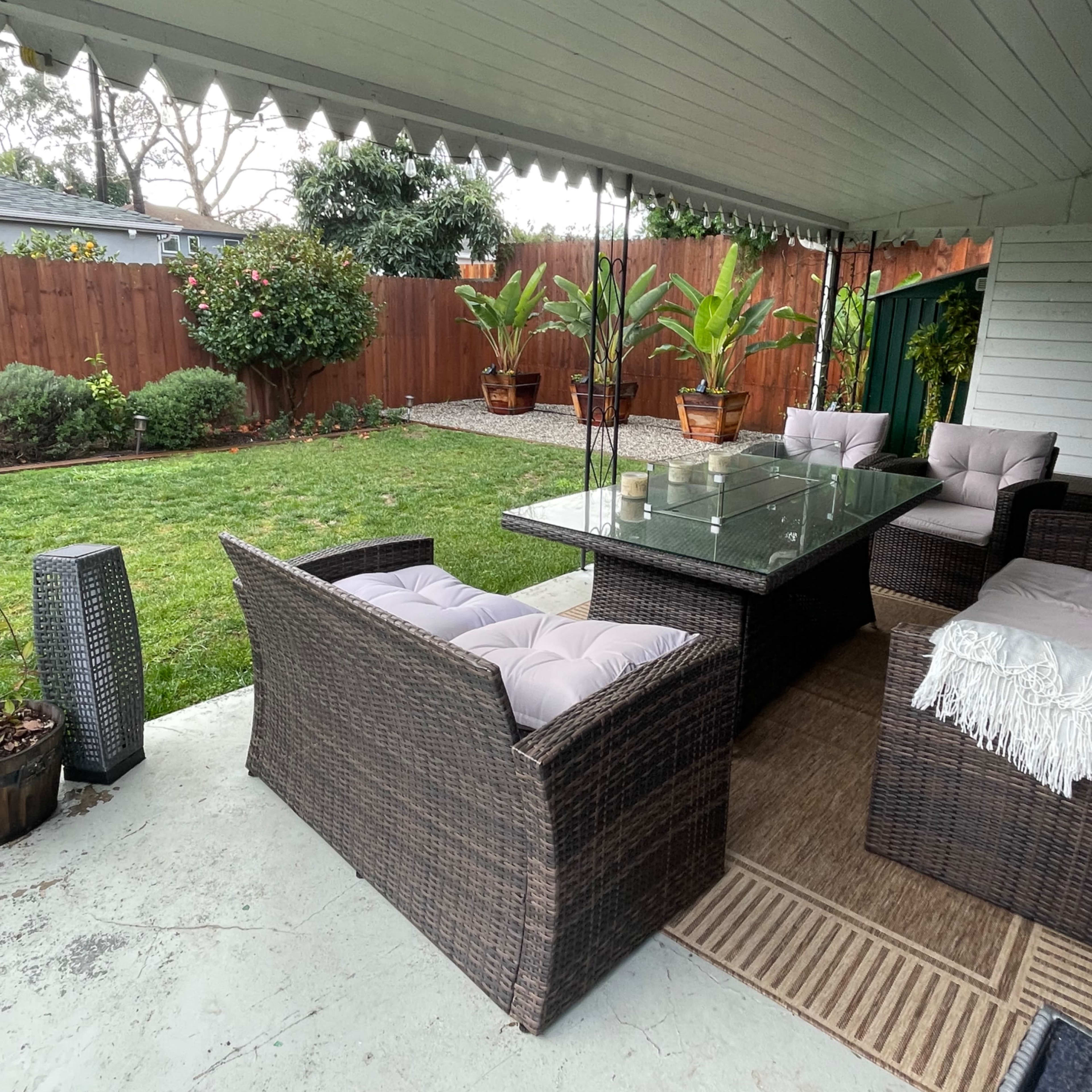 A covered patio features wicker furniture, a glass table, and planters with plants, overlooking a grassy backyard.