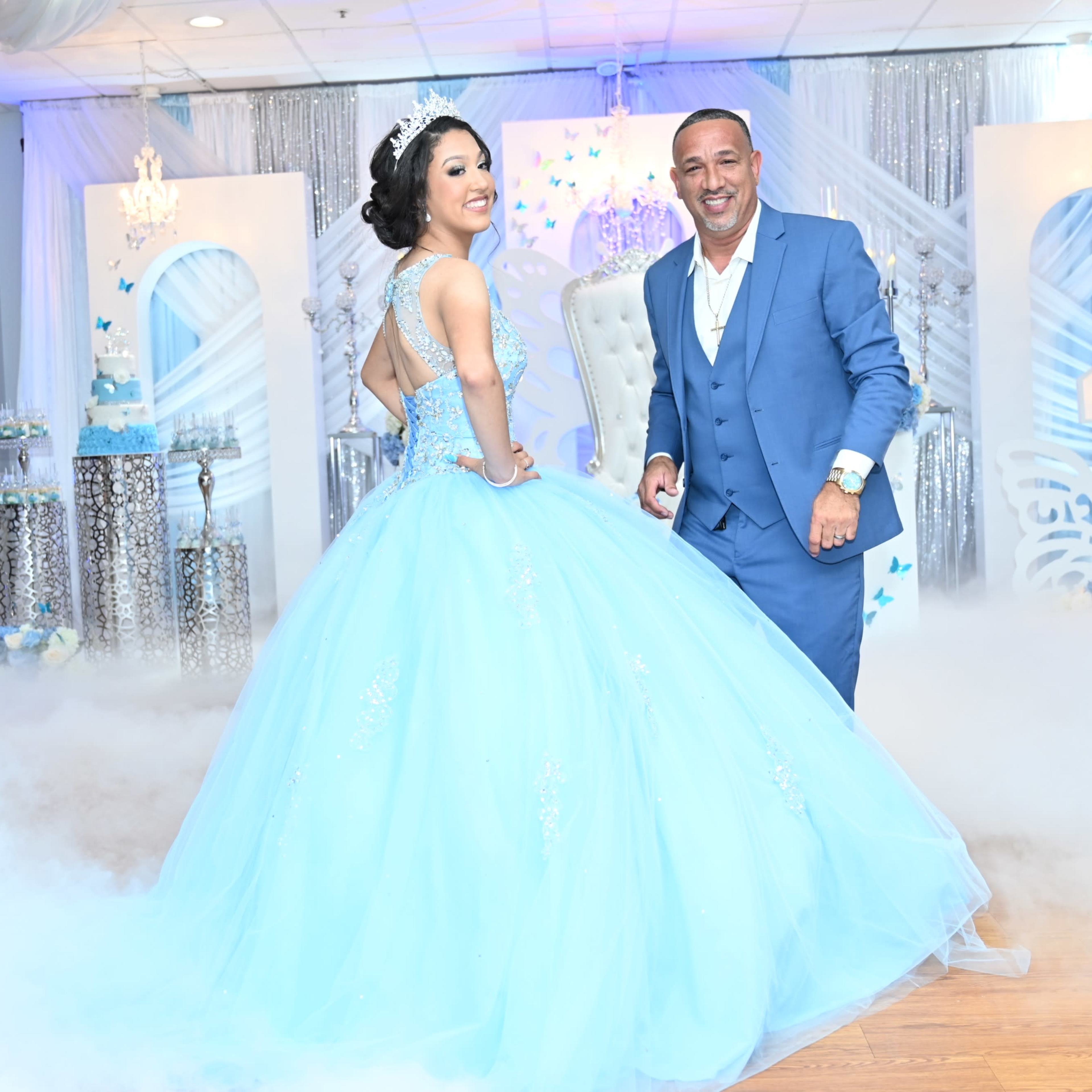 A young woman in a light blue ball gown poses with a man in a blue suit at a decorated event space with a misty floor.
