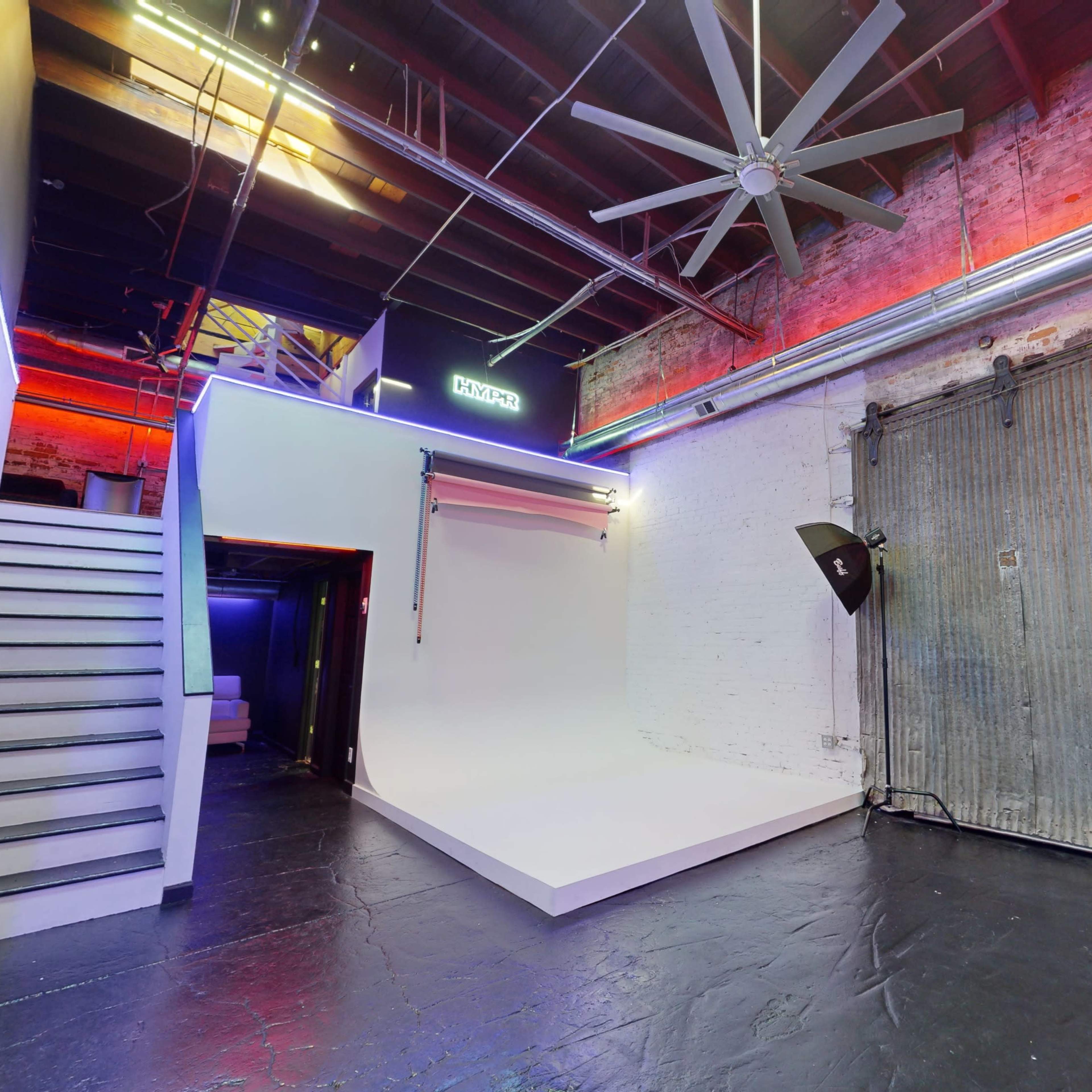 A spacious studio with a white backdrop, large ceiling fan, and industrial-style brick walls, along with a set of stairs leading to an upper level.