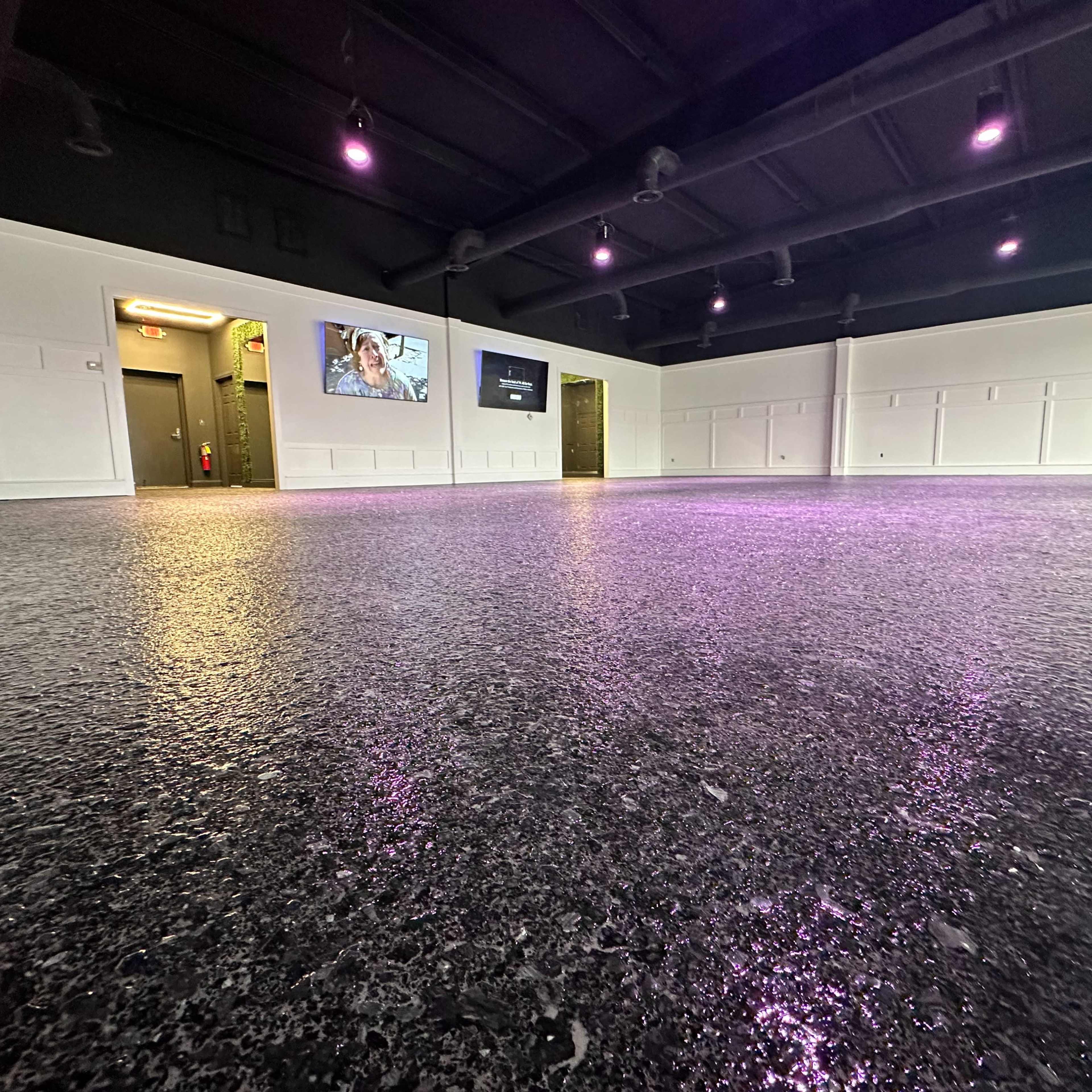 A spacious, brightly lit room with a polished floor, featuring purple-accented lighting and several doors leading to other areas.