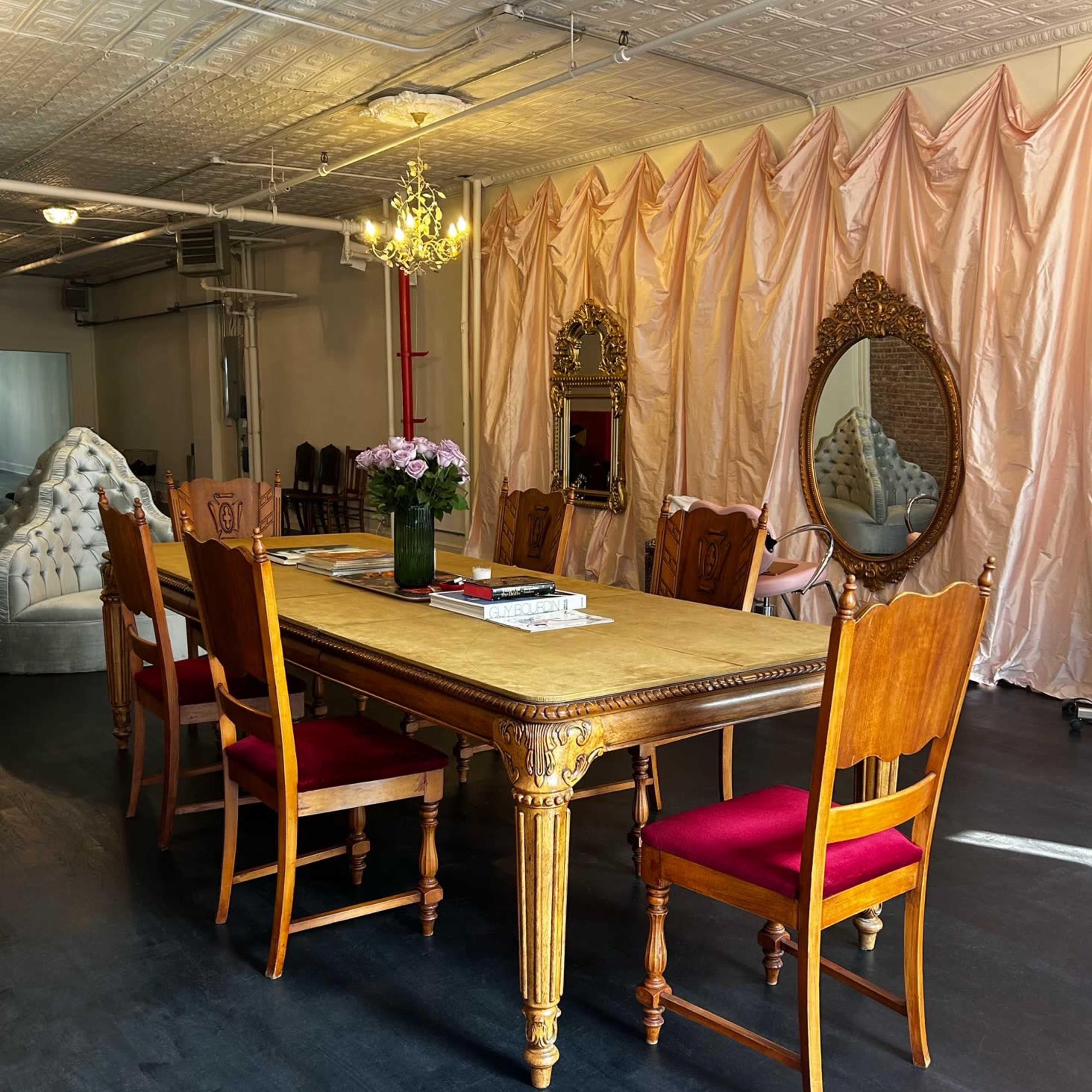 A long wooden dining table is surrounded by eight chairs in a well-lit room with mirrored accents and a fabric drape in the background.